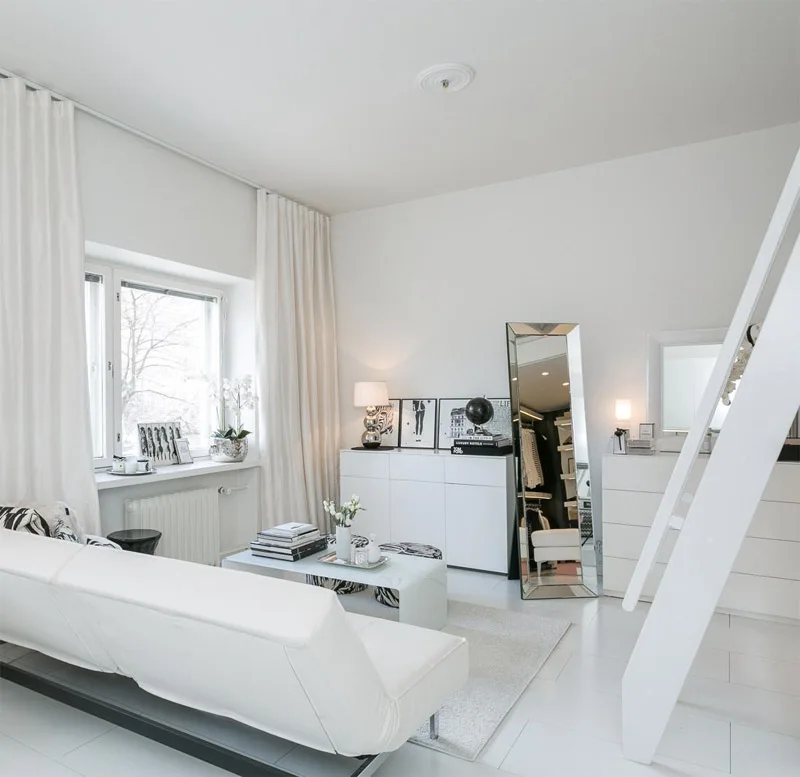 A bright Scandinavian studio living room with a white sofa, floor-to-ceiling curtains, a tilted floor mirror, and a white loft ladder.