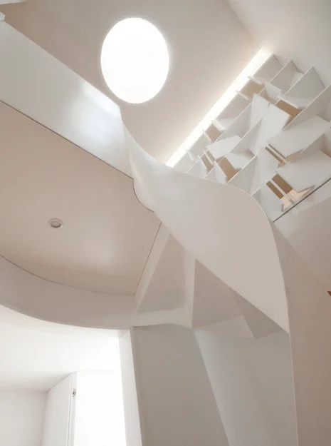 A sculptural curved white staircase ascends toward a circular skylight and a geometric built-in bookcase under a bright white ceiling.