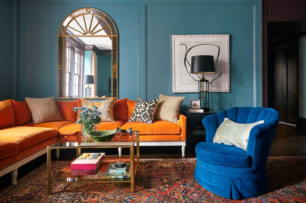 An eclectic living room with deep teal paneled walls, a vibrant orange sectional sofa, an arched gold-framed mirror, and a blue velvet armchair.