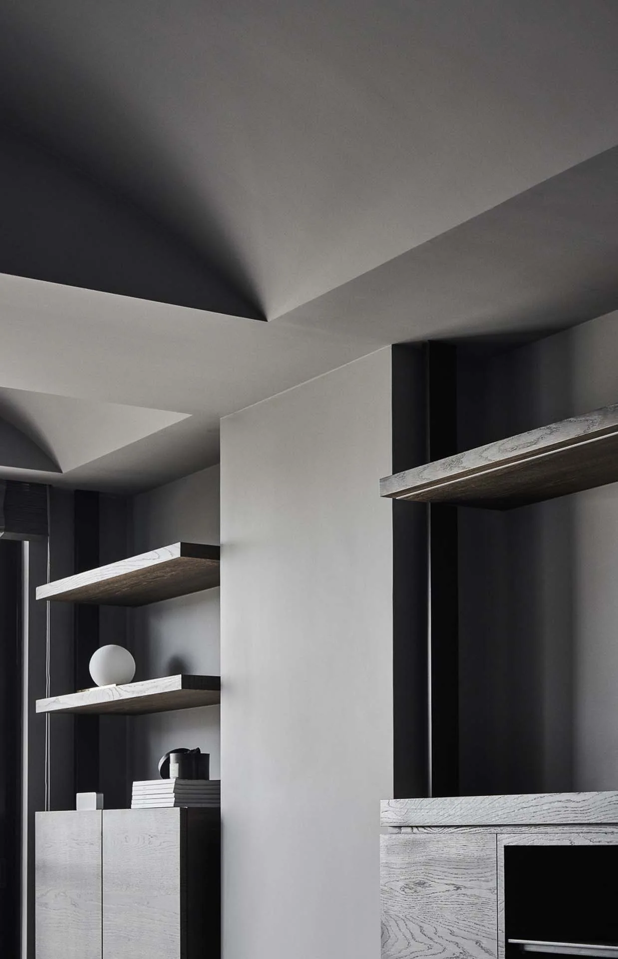 A minimalist grey room featuring angled ceiling architecture, textured dark wood floating shelves, and low cabinetry with minimal decor.