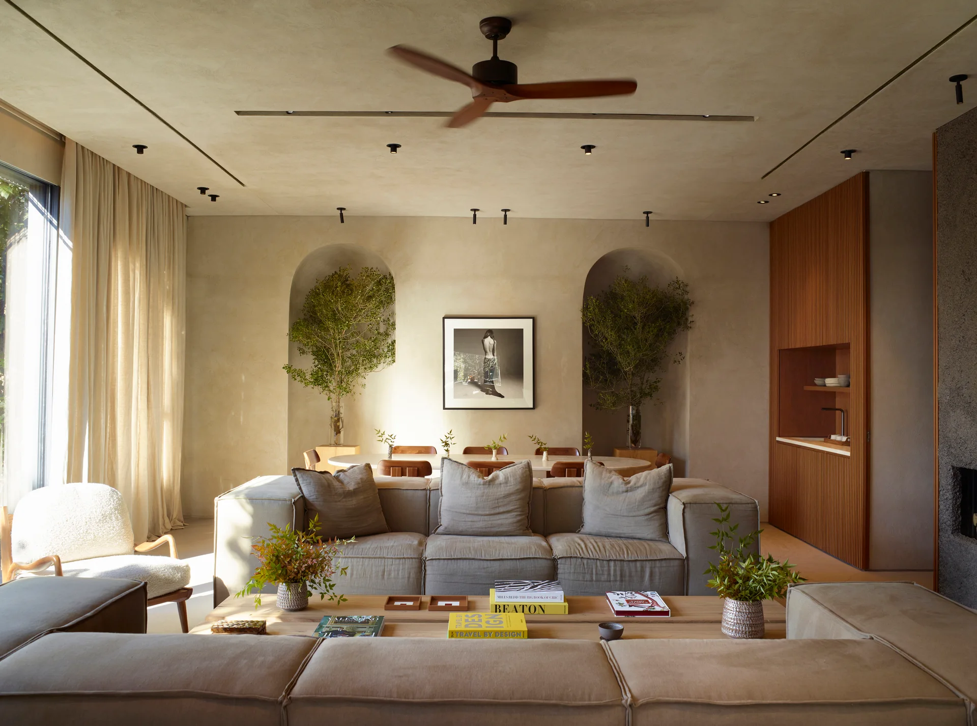 A warm neutral living room features a modular beige sofa, a low wood coffee table, and two arched plaster wall niches filled with leafy branches.