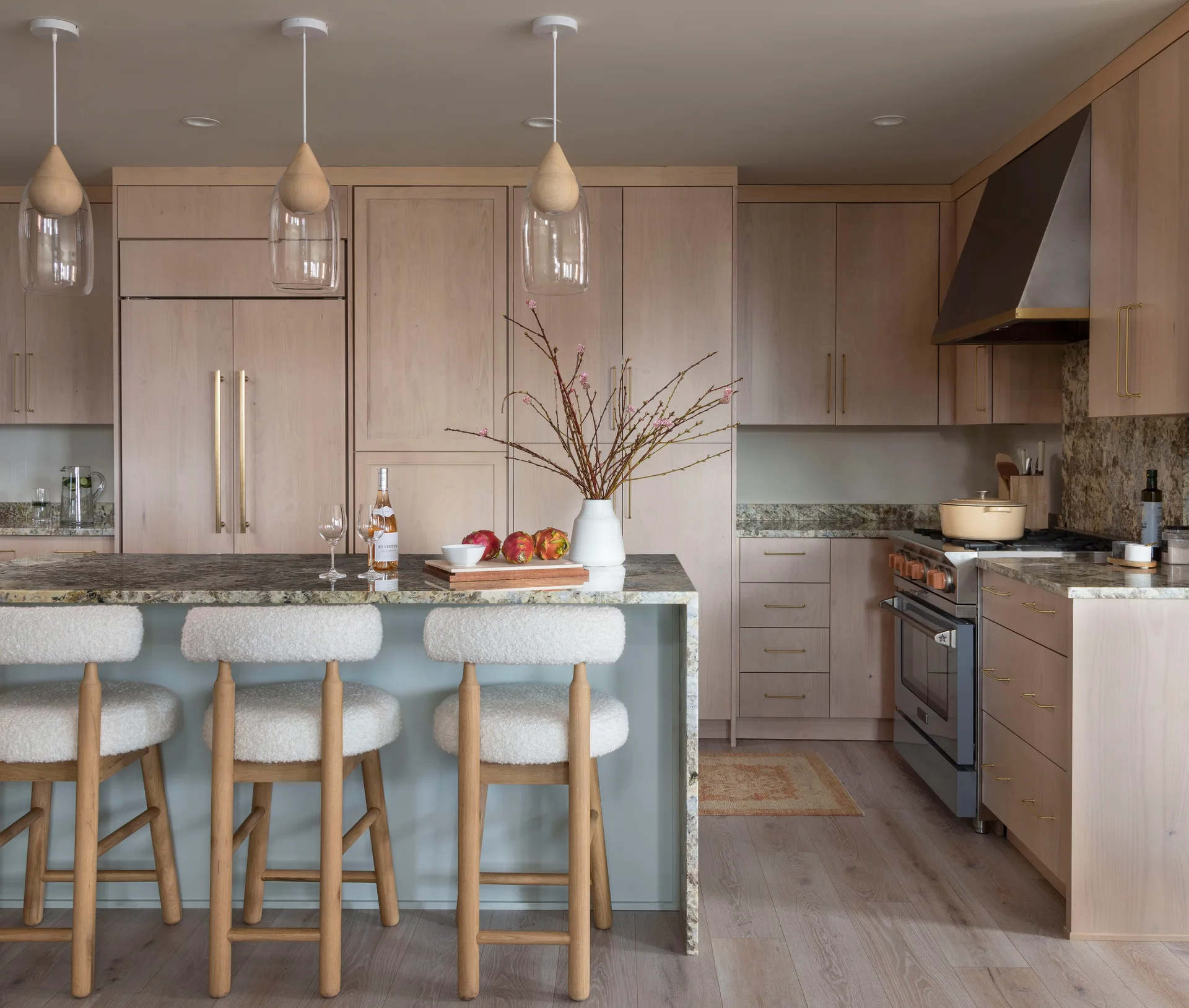 A modern kitchen features light wood cabinets, three clear glass pendant lights, and a stone-topped waterfall island with boucle bar stools.