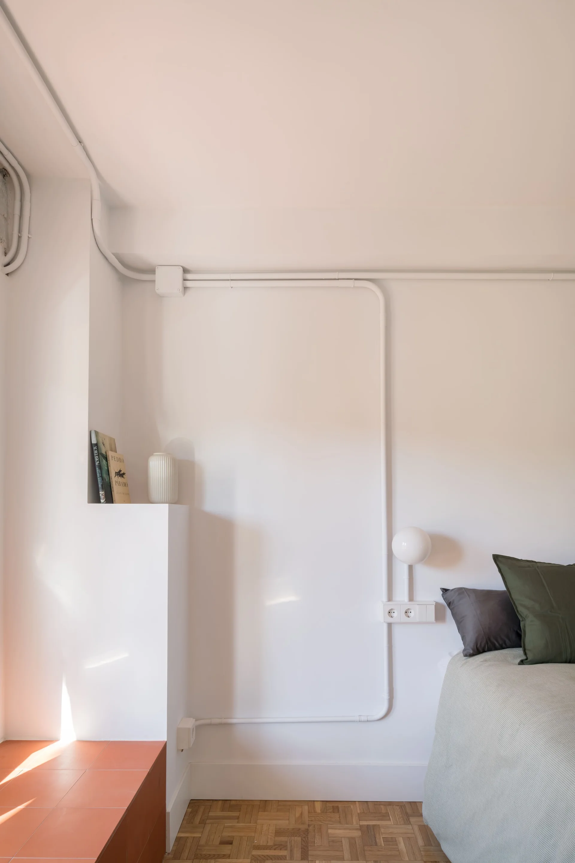 A minimalist bedroom corner features a bed with olive green pillows next to a white wall with exposed electrical conduit and a stepped built-in ledge.