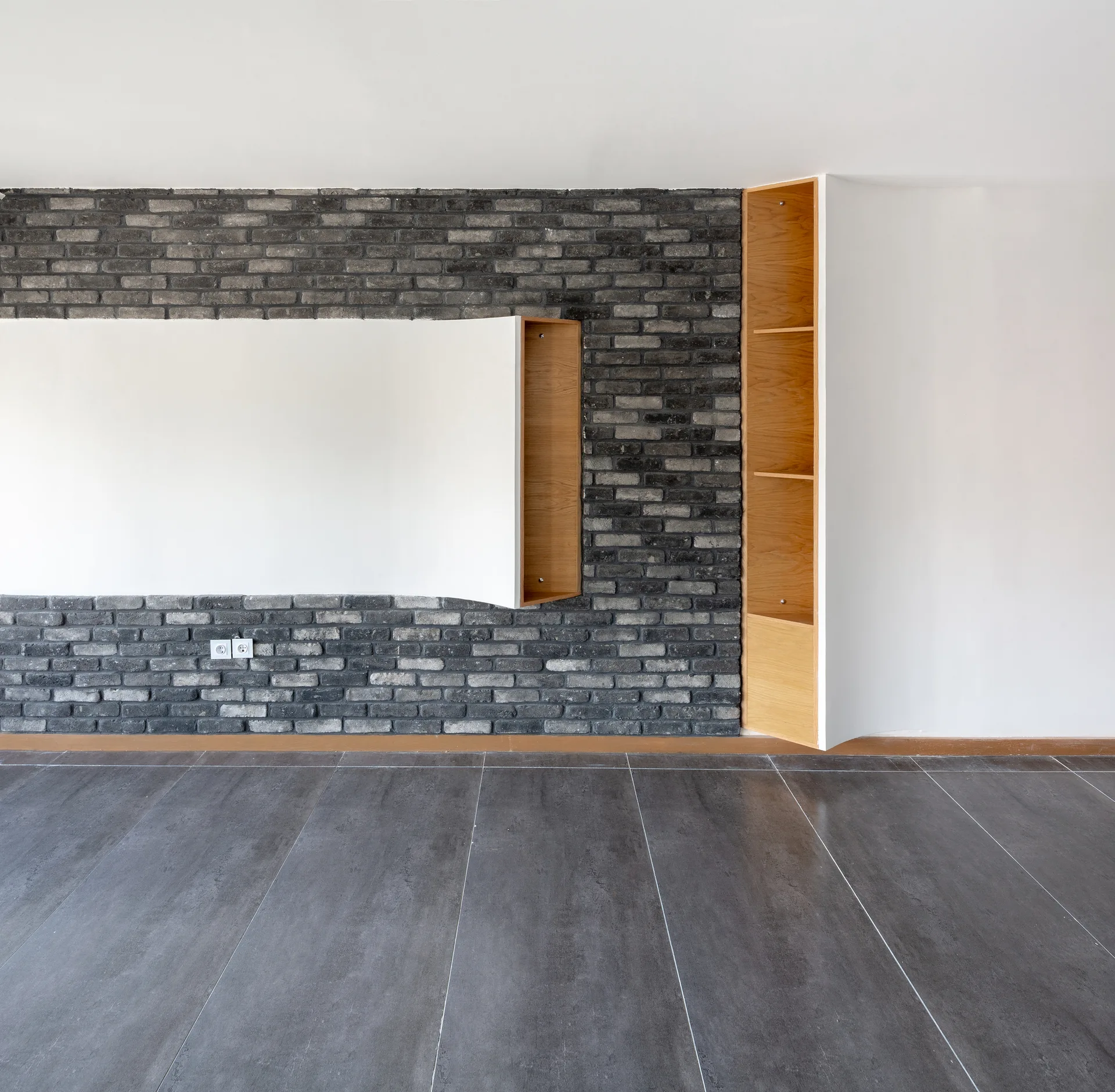 An empty room features an industrial dark brick accent wall with custom wooden shelving recesses and large gray floor tiles.