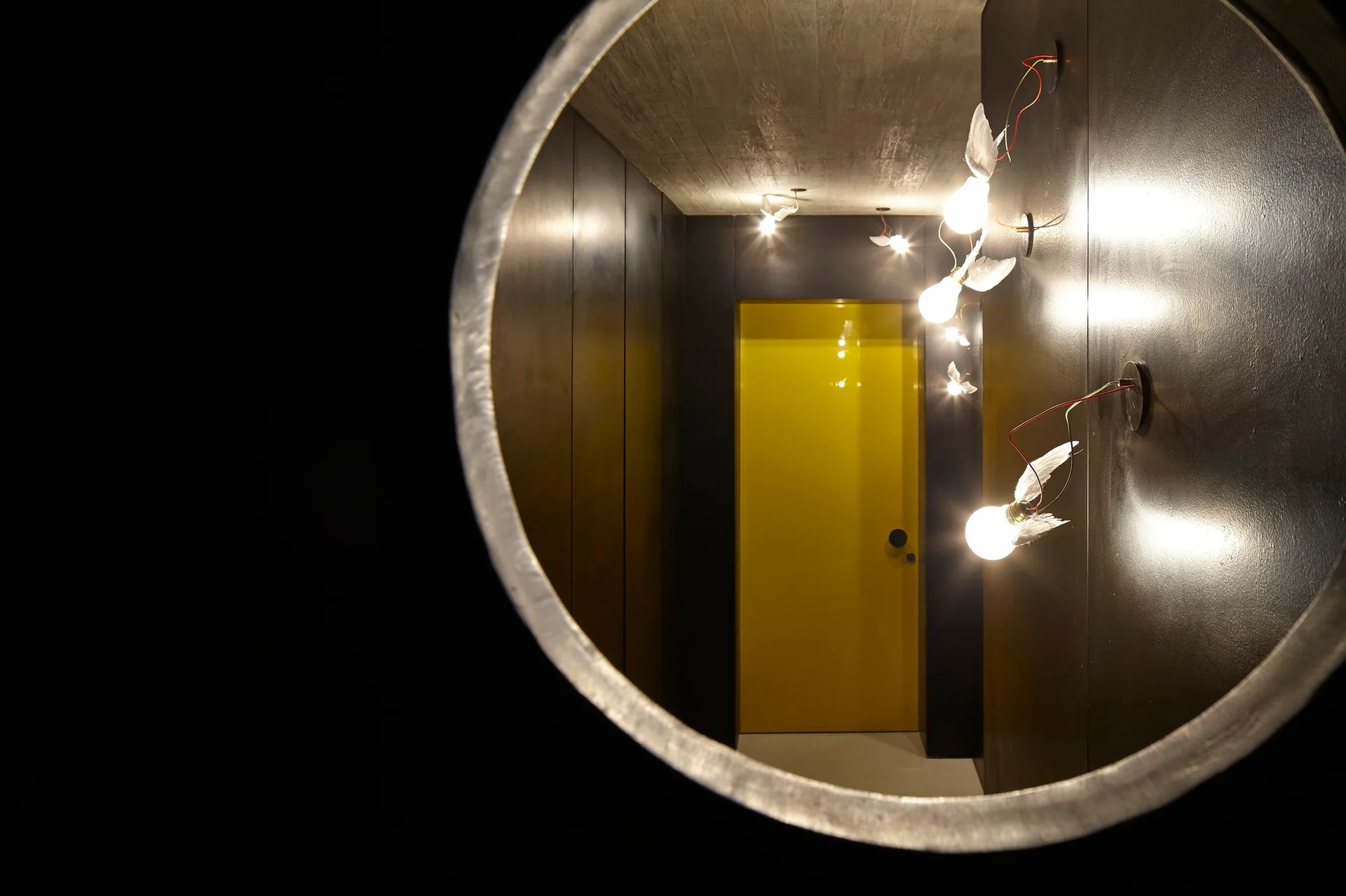 A dark hallway with black walls, an exposed concrete ceiling, and winged light fixtures leads to a bright yellow door, viewed through a circular frame.