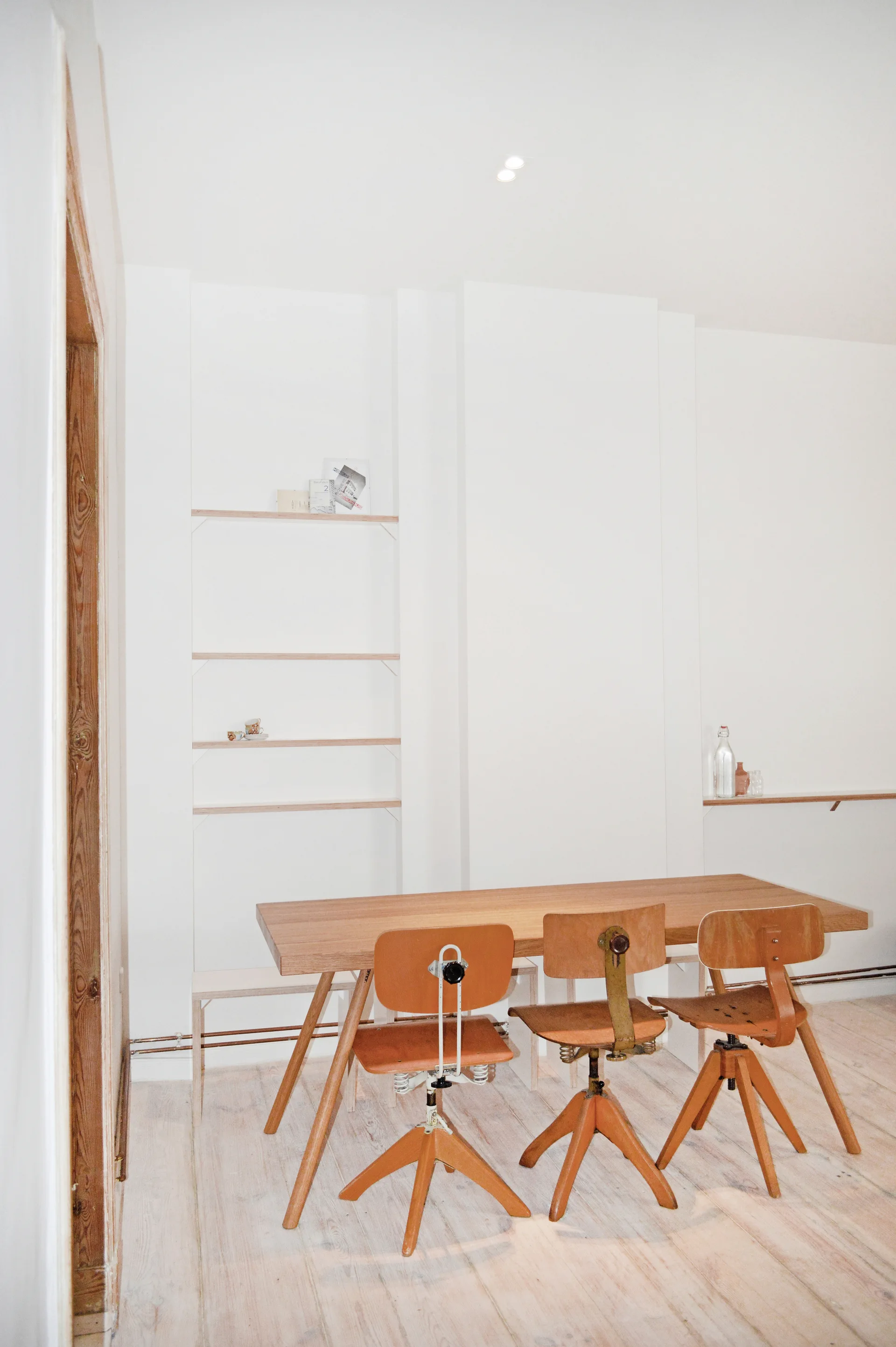 A light wood dining table and three vintage wooden swivel chairs sit against a plain white wall with recessed shelves.