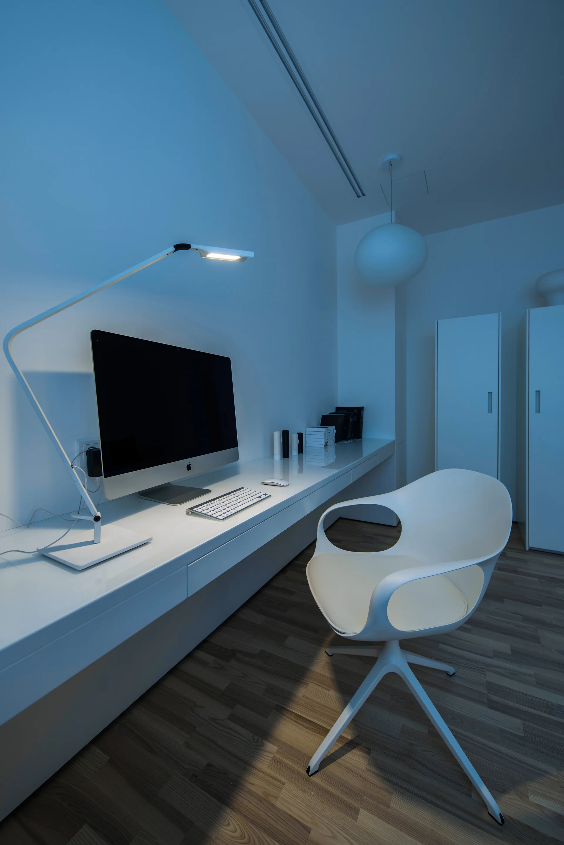 A modern home office features a long white floating desk with a computer, a sculptural white swivel chair, and wood plank flooring.
