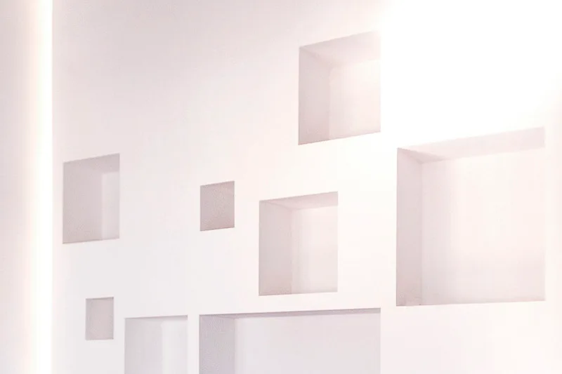 A smooth white plaster wall features several recessed square niches of varying sizes in an asymmetrical configuration.
