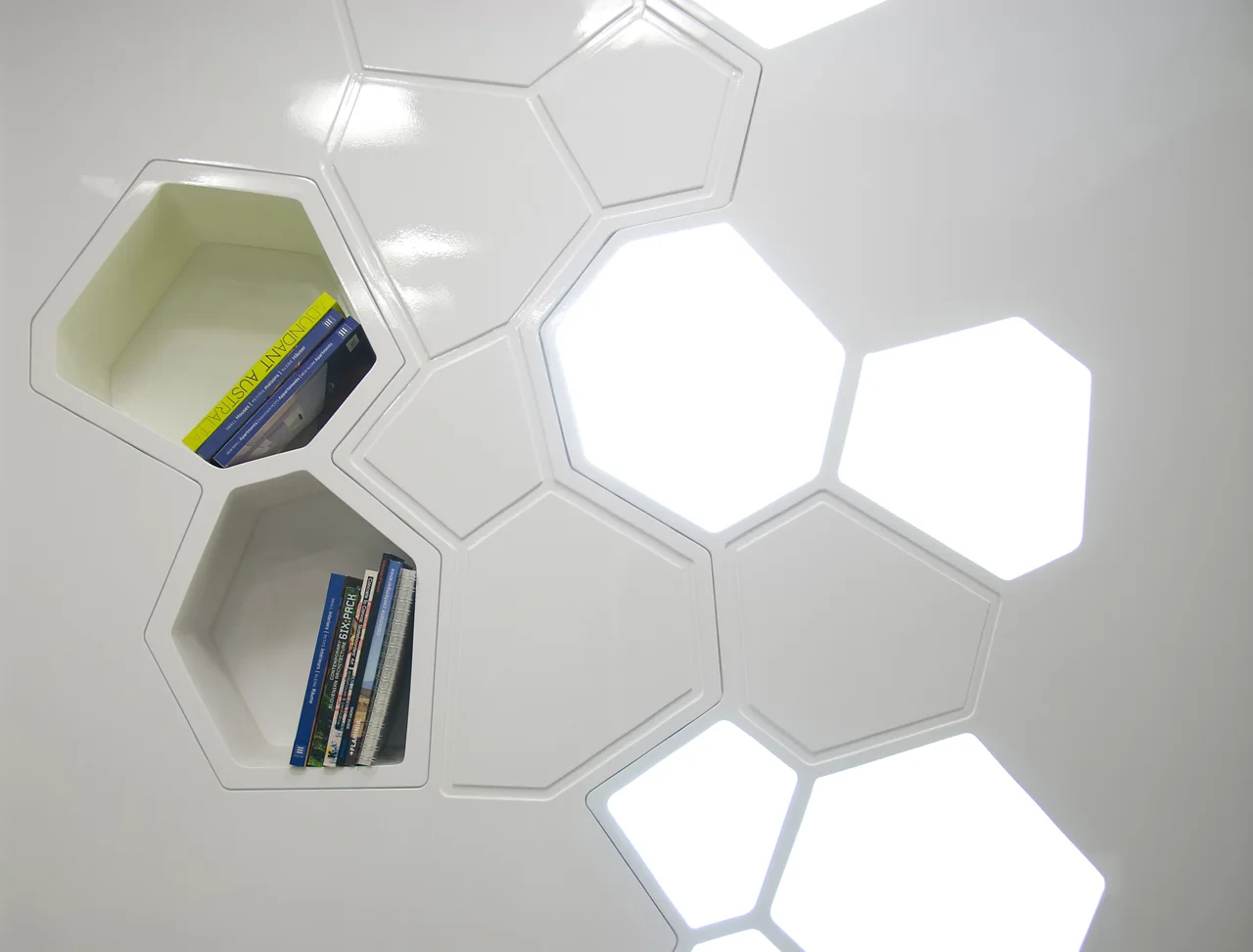 A high-gloss white wall features integrated hexagonal paneling with glowing backlit panels and recessed storage cubbies containing books.