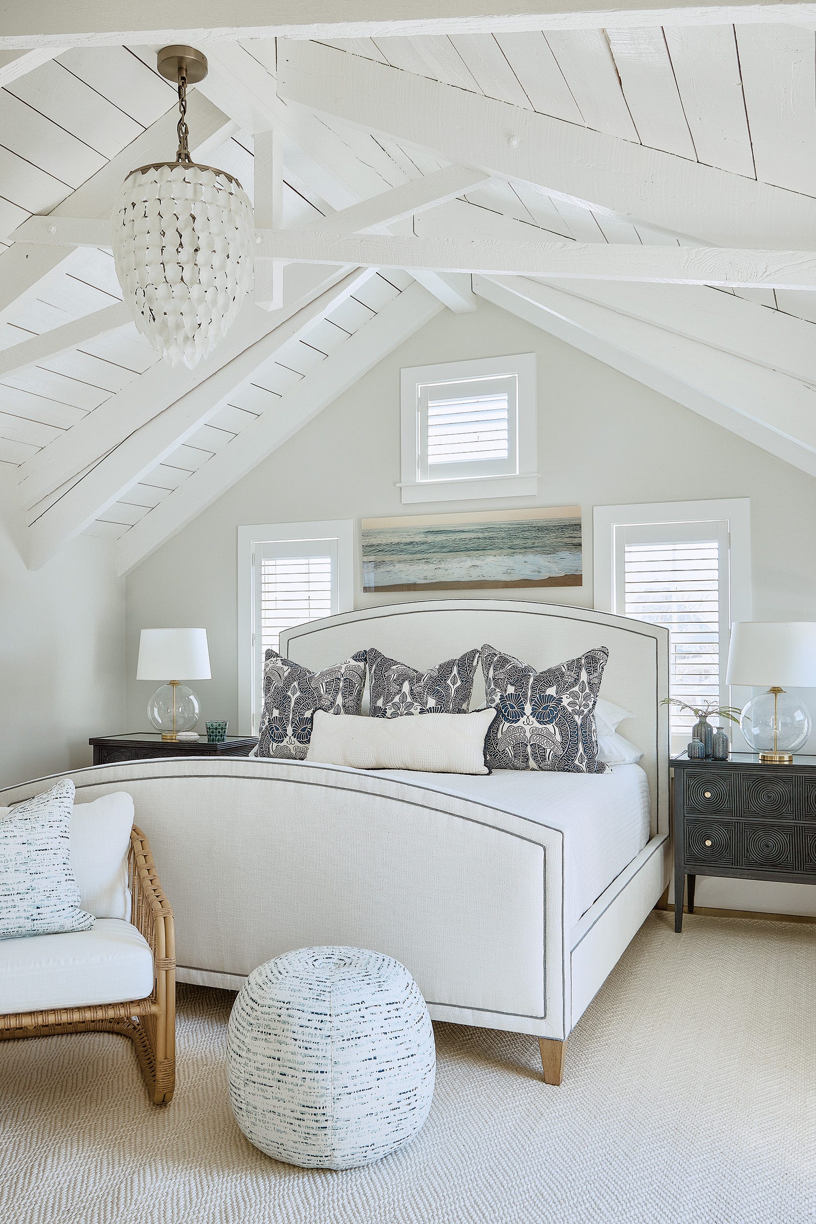 A bright coastal bedroom features an upholstered white bed beneath a vaulted planked ceiling, flanked by dark nightstands and glass lamps.