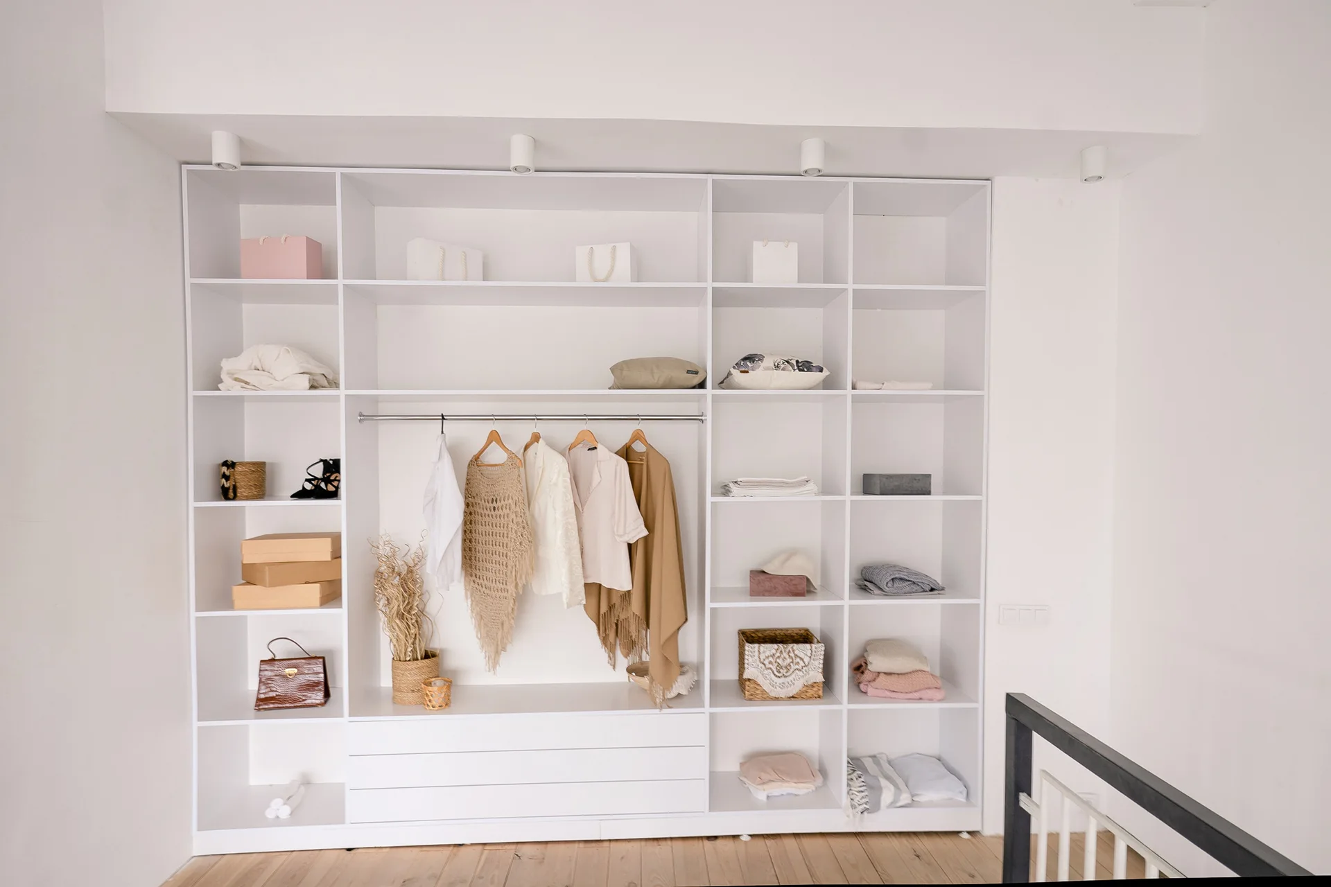 An open floor-to-ceiling white wardrobe system features grid shelving, a central hanging rod, and neatly arranged clothing and neutral accessories.