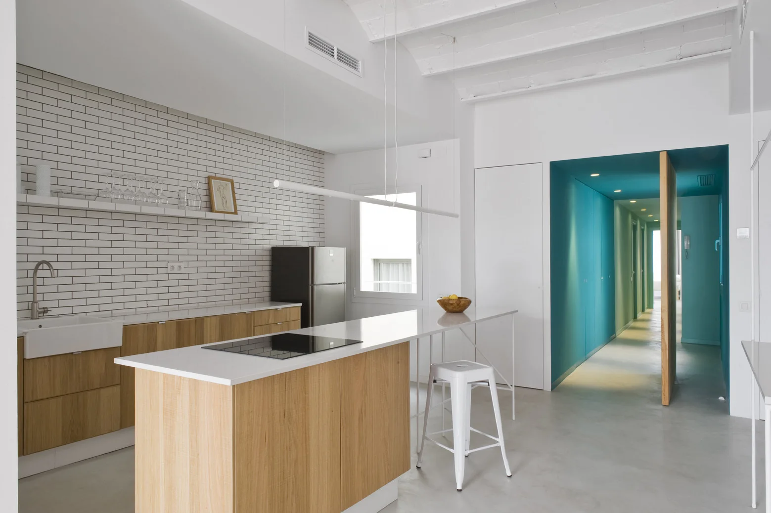 A modern kitchen with natural wood cabinetry, a white brick tile backsplash, a long island counter, and a bright blue hallway in the background.