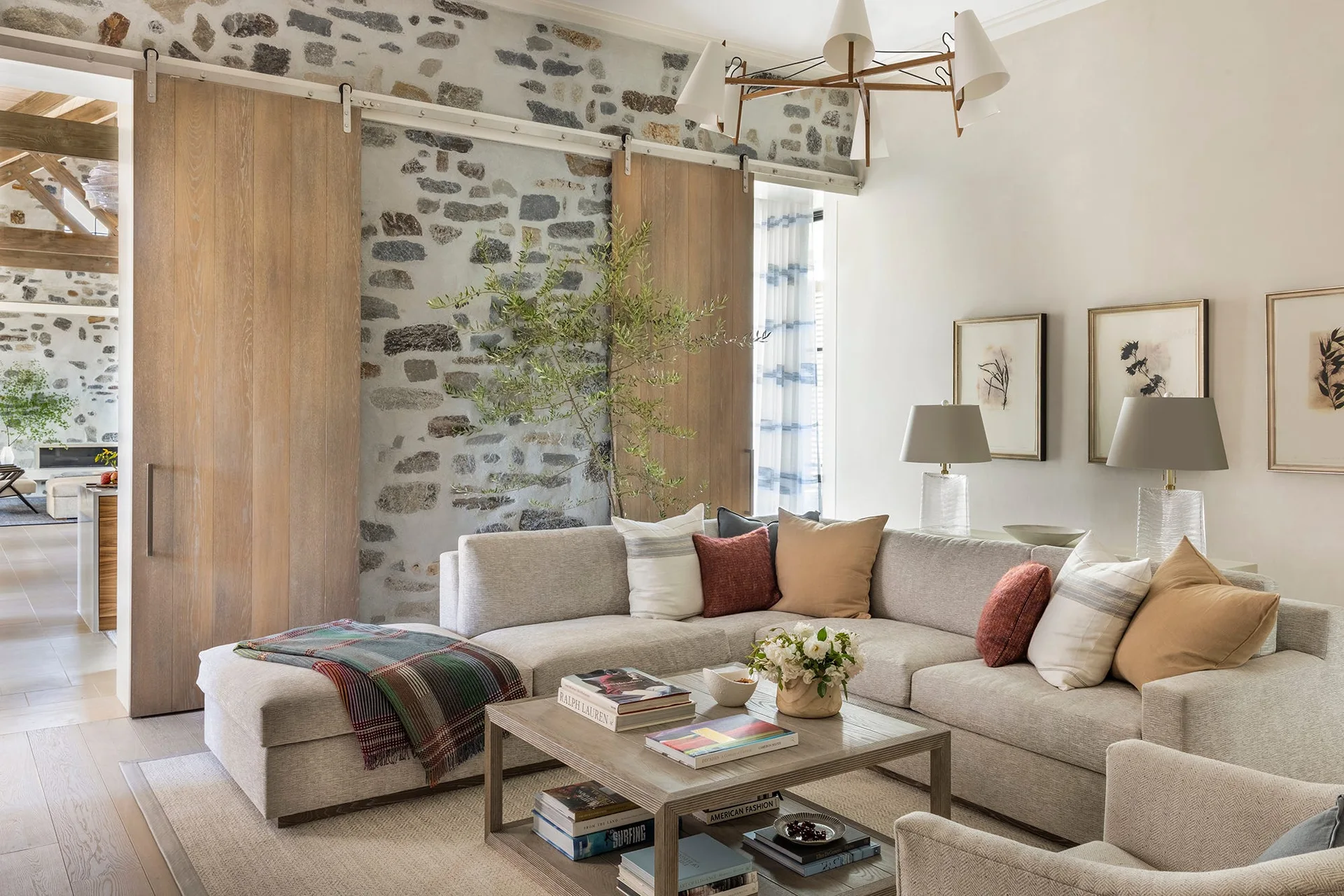A light grey sectional sofa and wooden coffee table sit in a living room with an exposed stone wall and sliding wood barn doors.