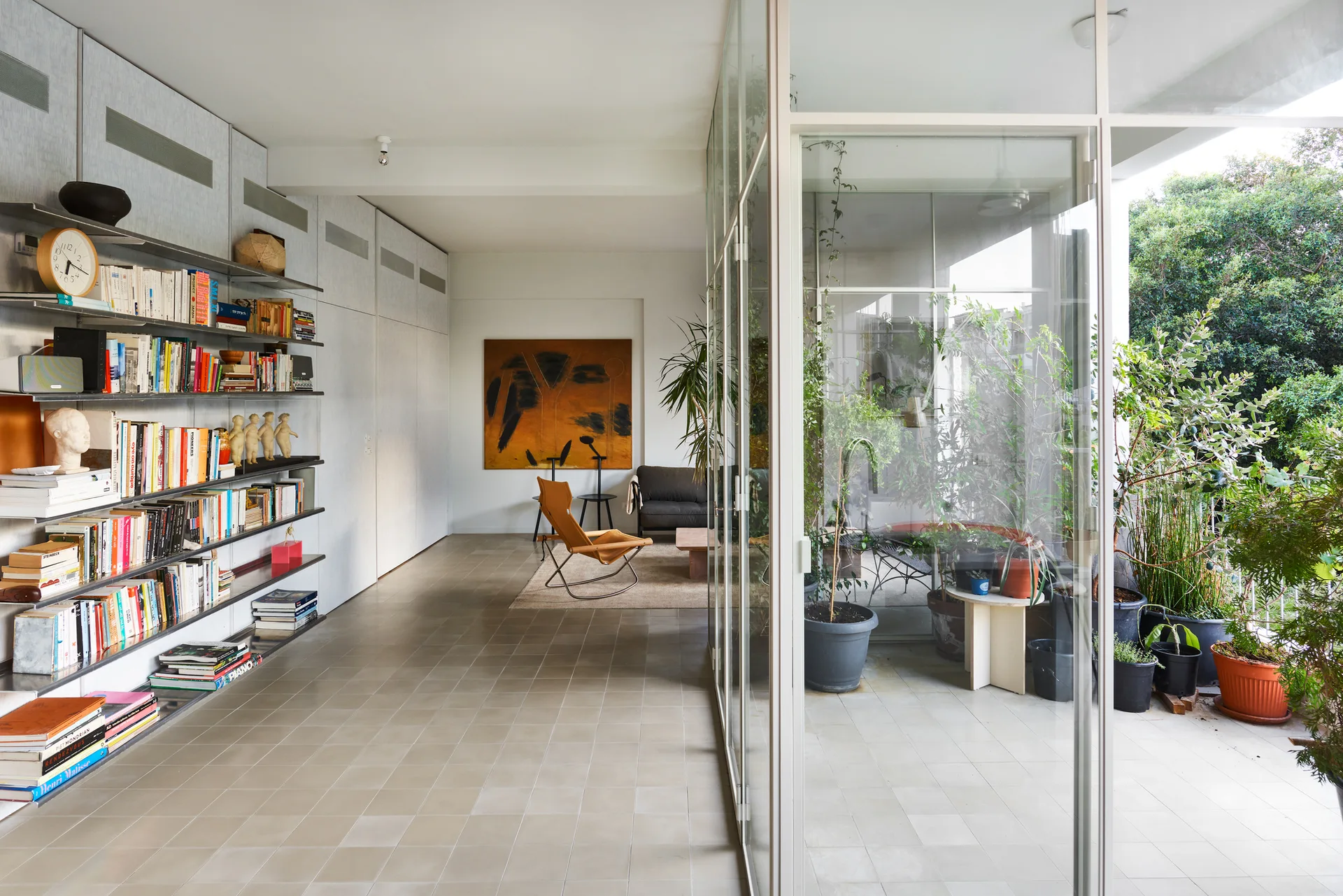 A modern living room features a long wall-mounted metal bookshelf, tile flooring, and a glass-enclosed balcony filled with green potted plants.