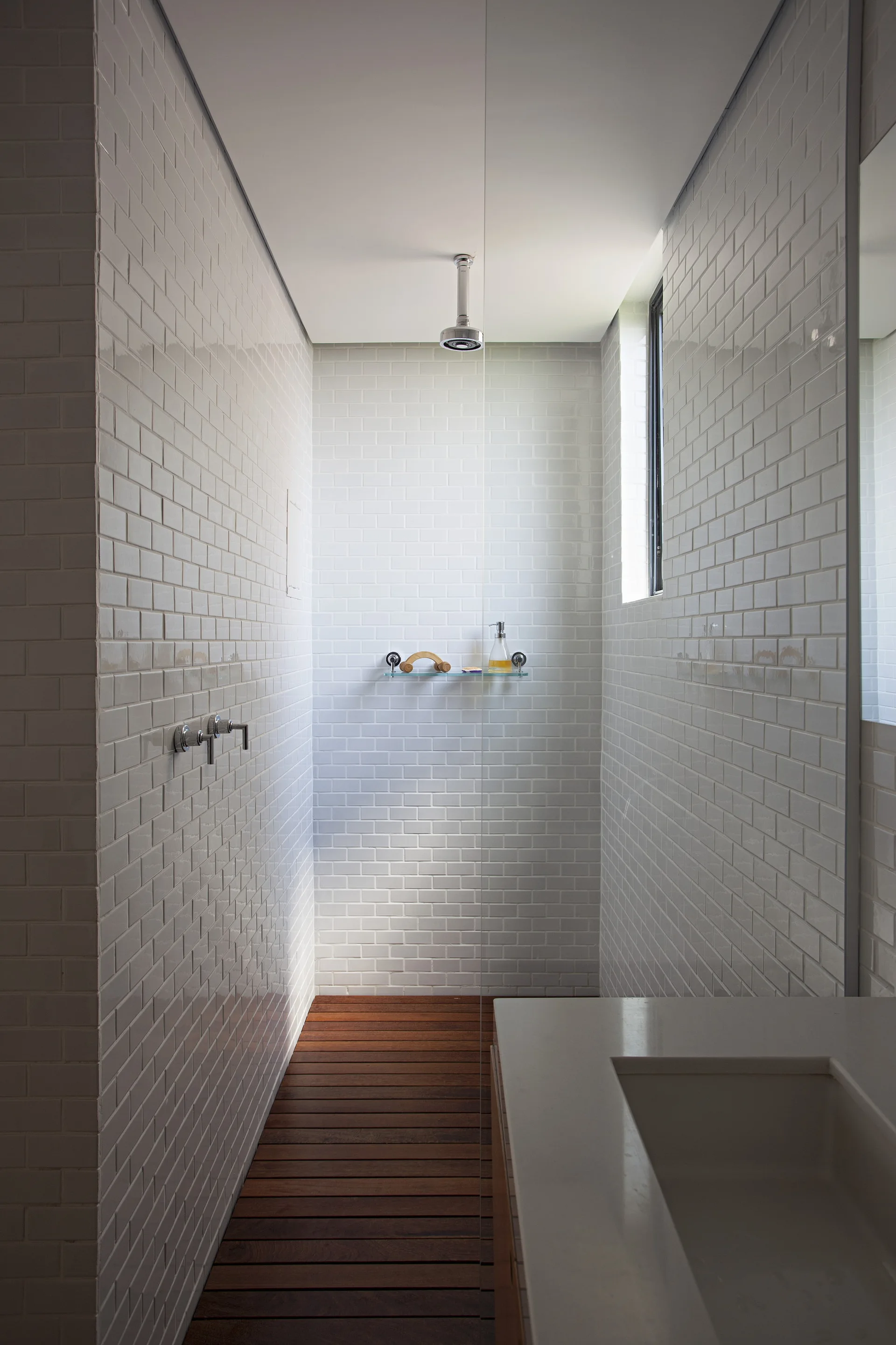 A modern walk-in shower featuring glossy white subway-tiled walls, a slatted wood plank floor, a ceiling-mounted showerhead, and a frameless glass partition.