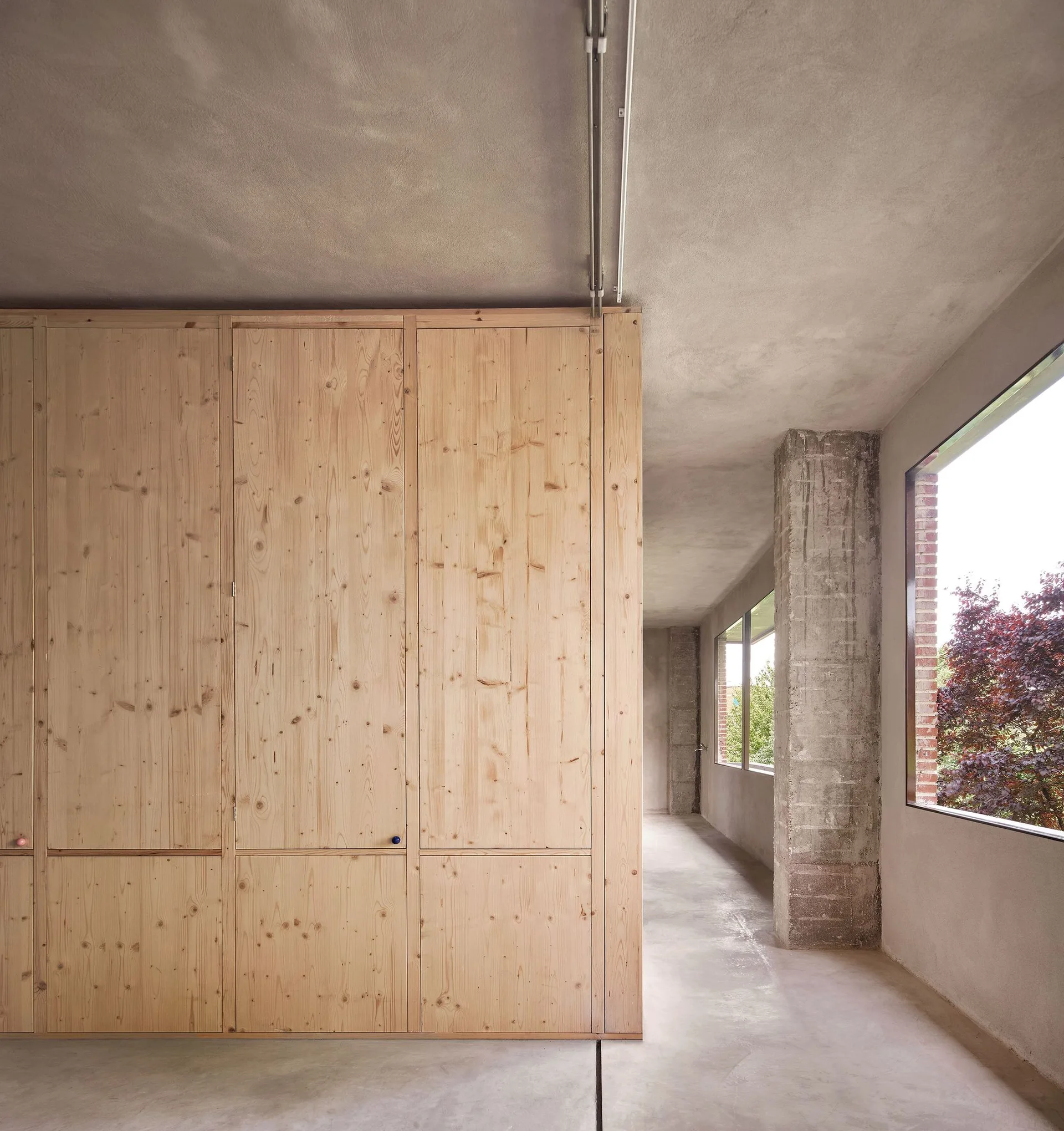 A spacious minimalist hallway features a tall wall of knotty pine wardrobes alongside raw concrete columns and a large window looking outdoors.