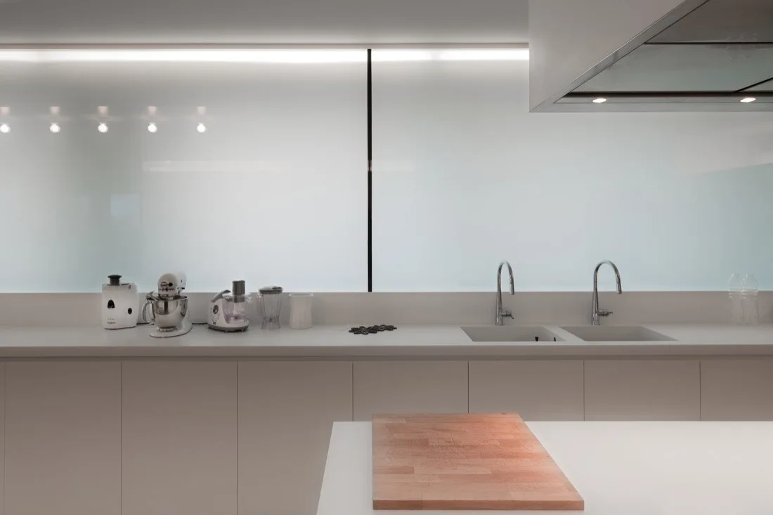 A minimalist white kitchen features a long countertop with double sinks, small appliances, and a large backlit frosted glass backsplash wall.