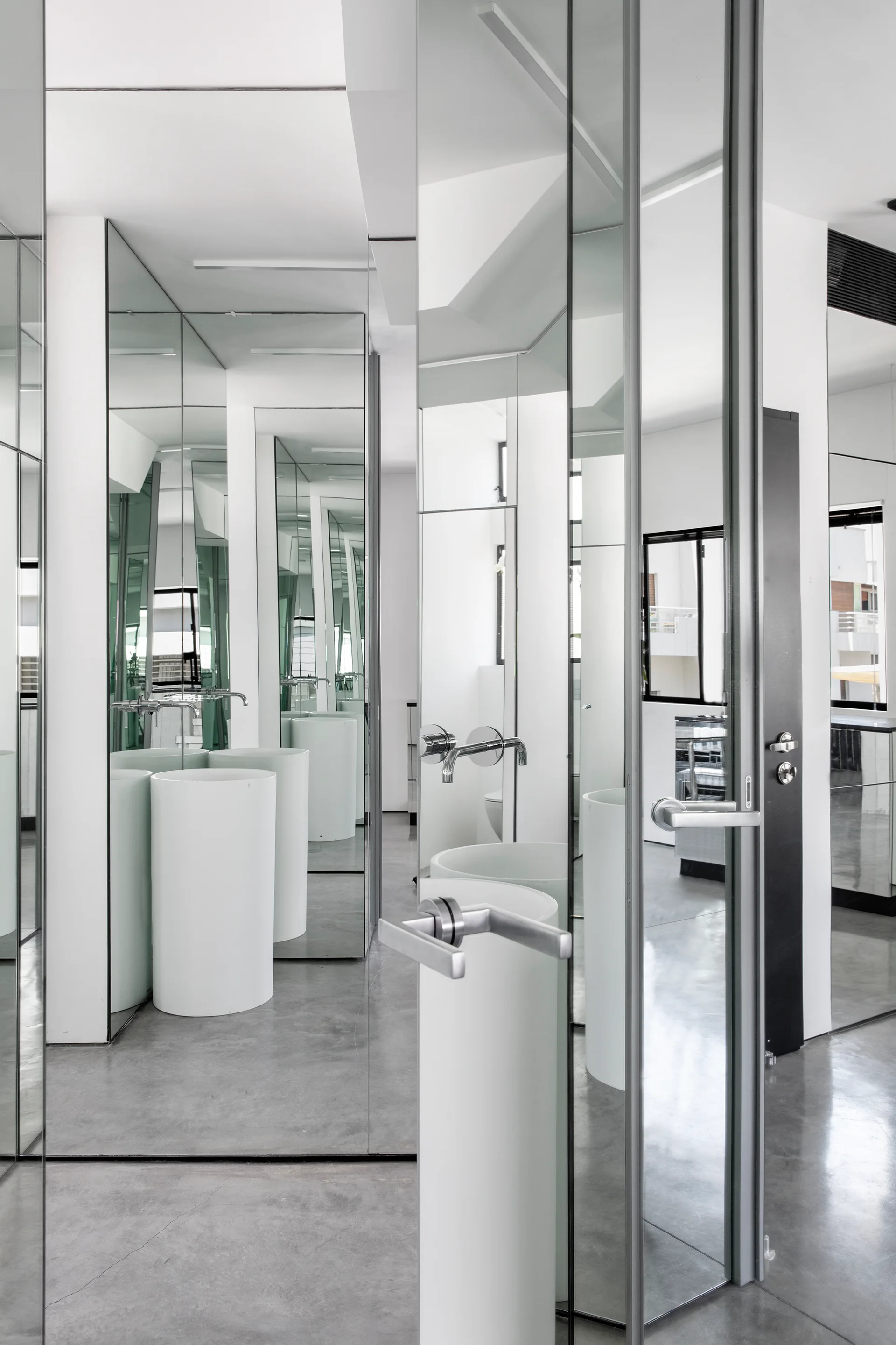 A minimalist bathroom features white cylindrical pedestal sinks and expansive floor-to-ceiling mirrored walls reflecting polished concrete floors.