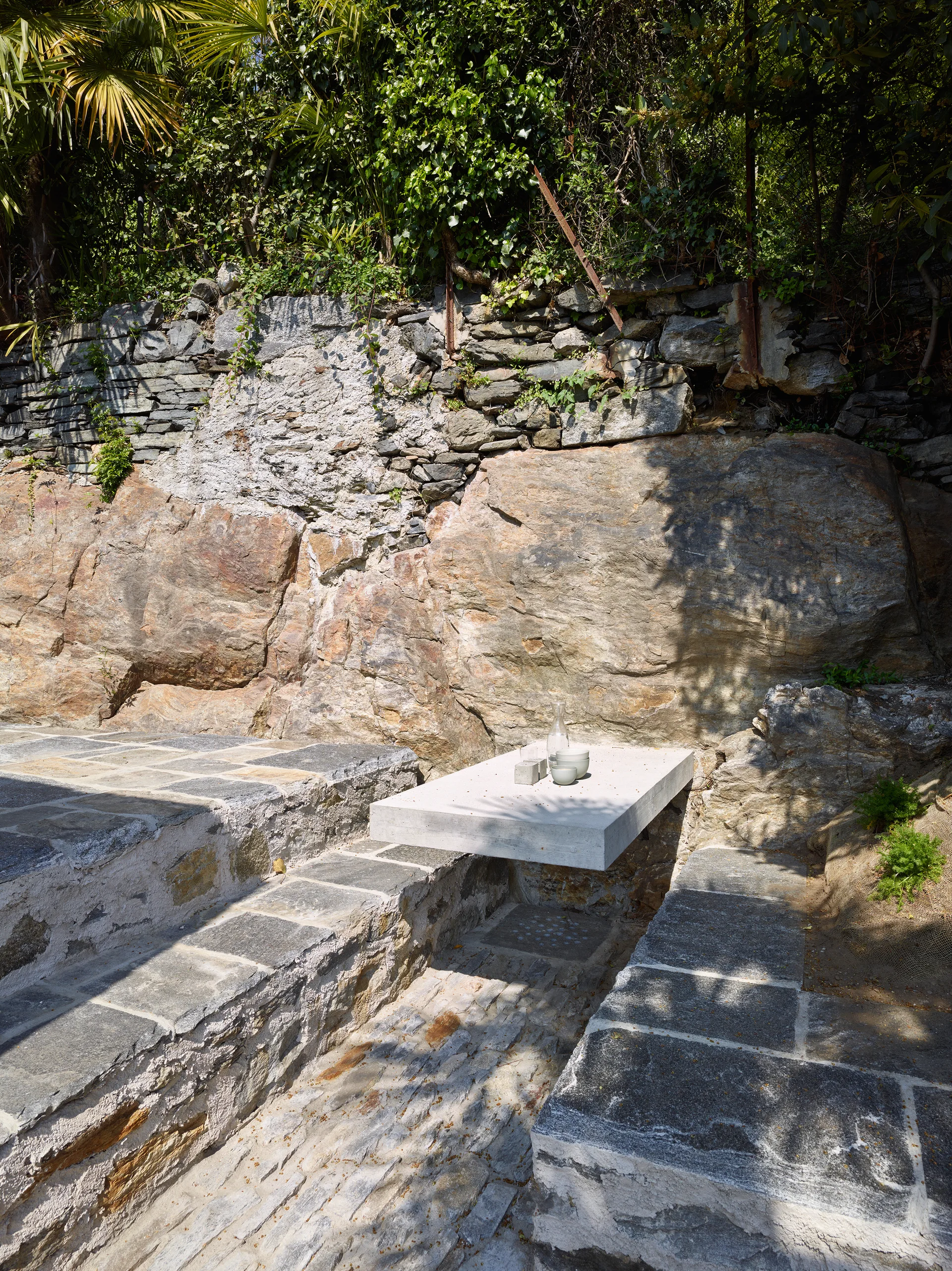 A smooth grey concrete table cantilevers over a stone paved patio next to a natural rugged rock wall.