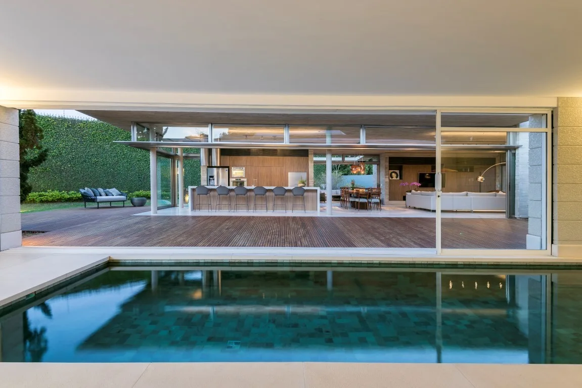 A sleek swimming pool overlooks a wooden deck and an open-concept kitchen and living area visible through large sliding glass doors.