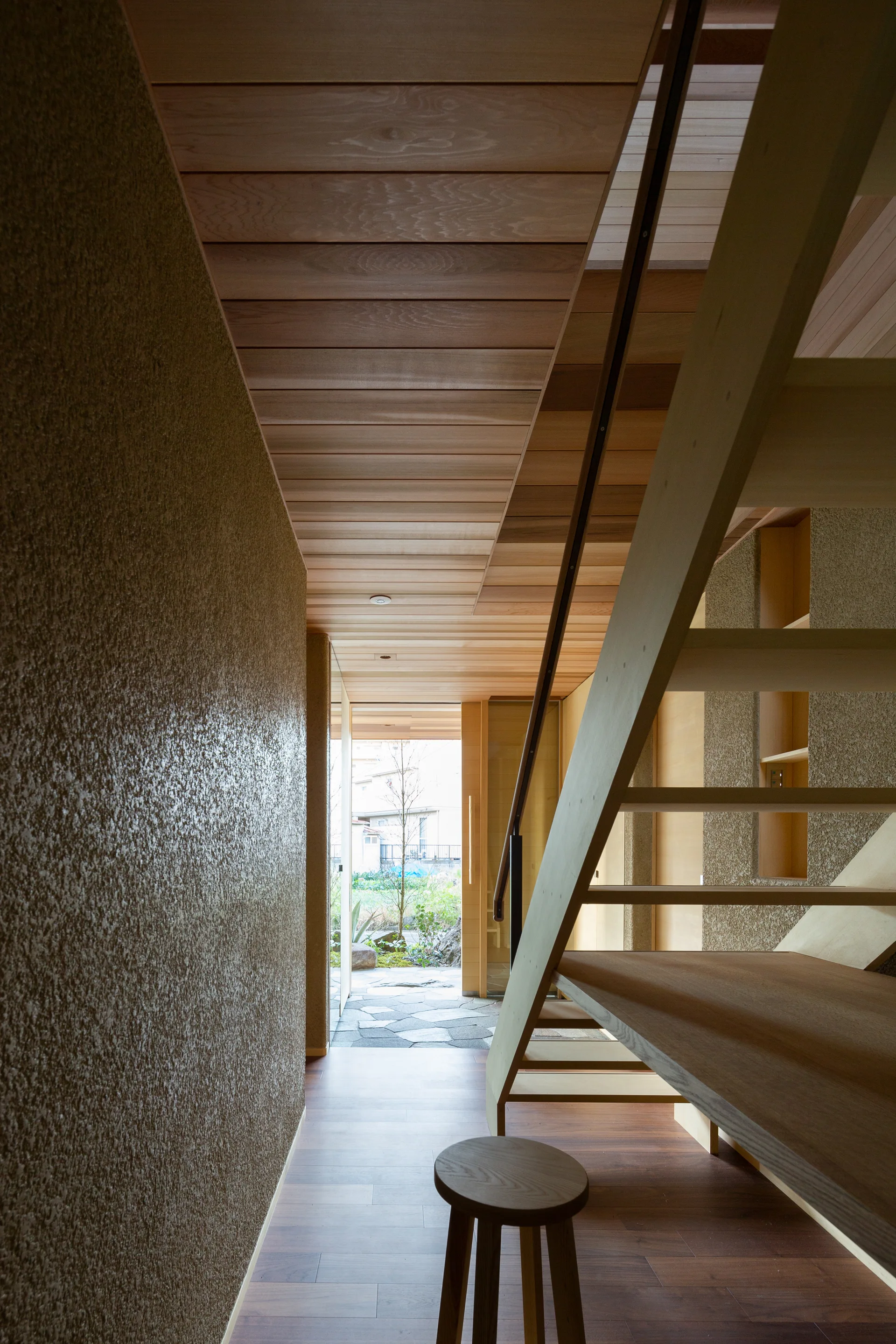 A narrow wood-paneled entryway features a minimalist open-tread staircase and a textured plaster wall leading to an exterior garden.
