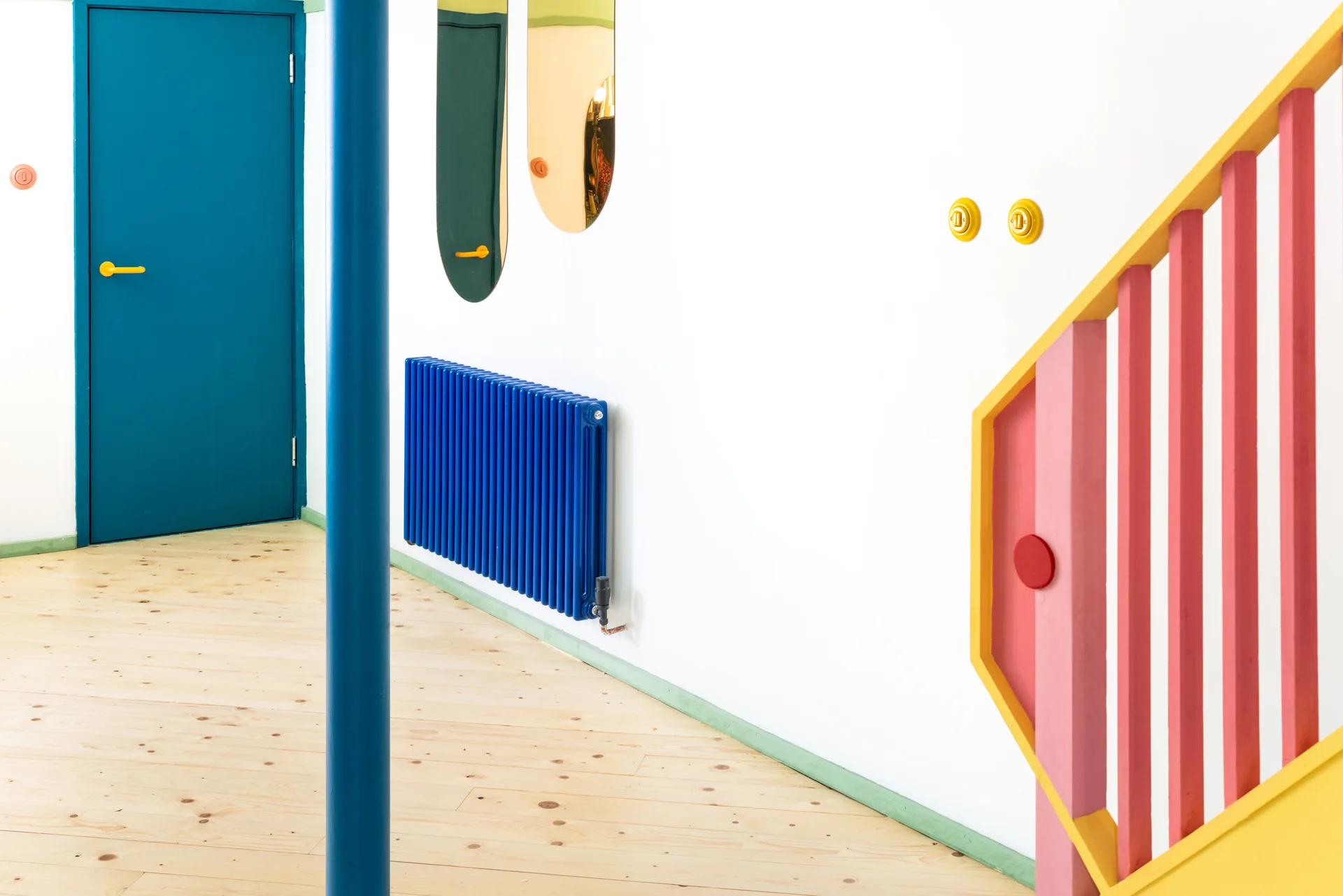 A bright white hallway featuring a bold blue door, a matching blue radiator, and a pink and yellow painted wooden staircase railing on light wood floors.
