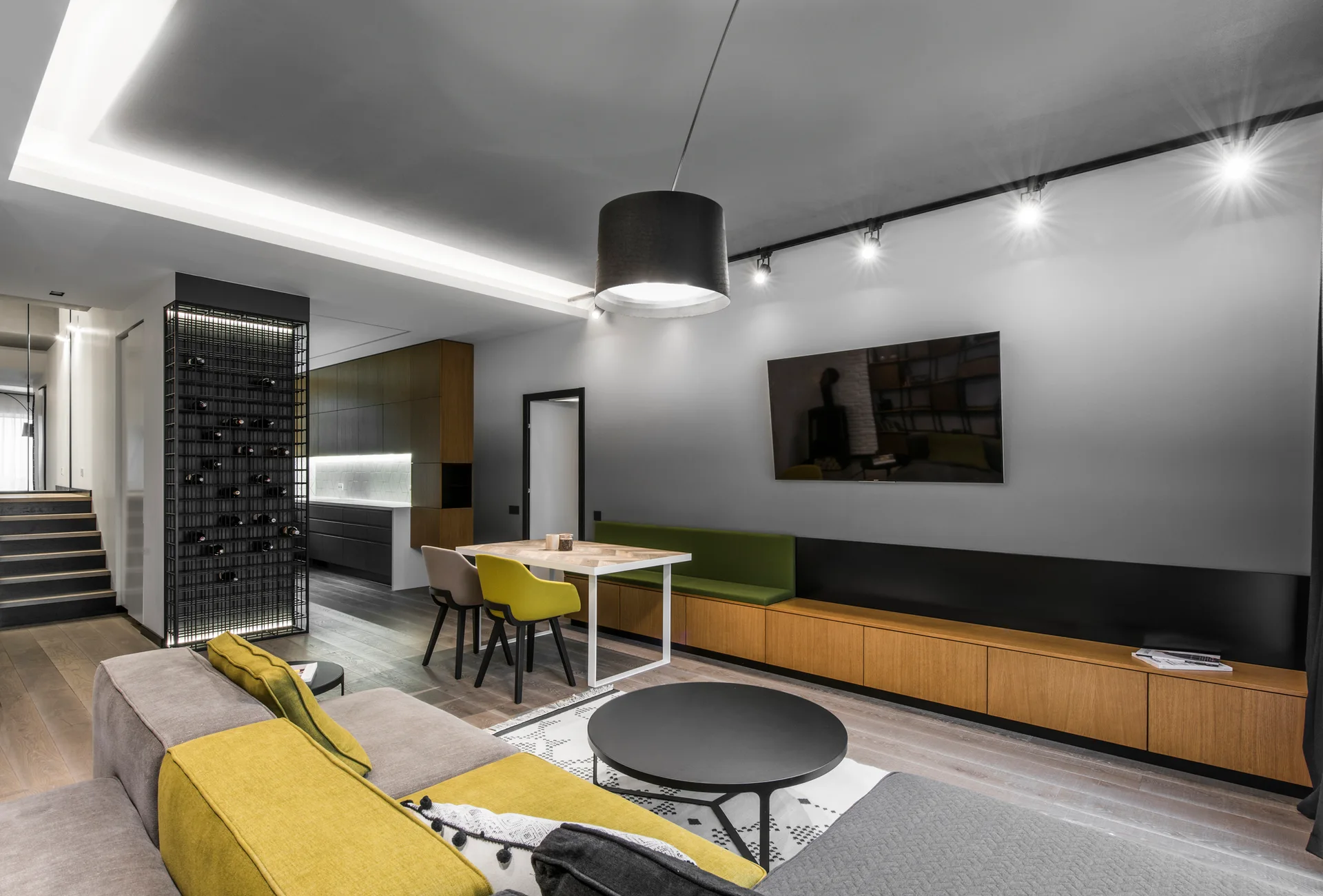 A contemporary living area features a grey modular sofa with yellow cushions, a built-in wood media console, and an integrated dining bench.
