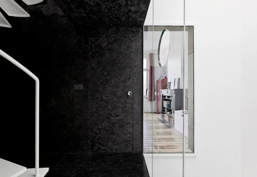 A dark hallway features a textured black OSB wall with a flush door next to a clear glass partition showing a bright room beyond.