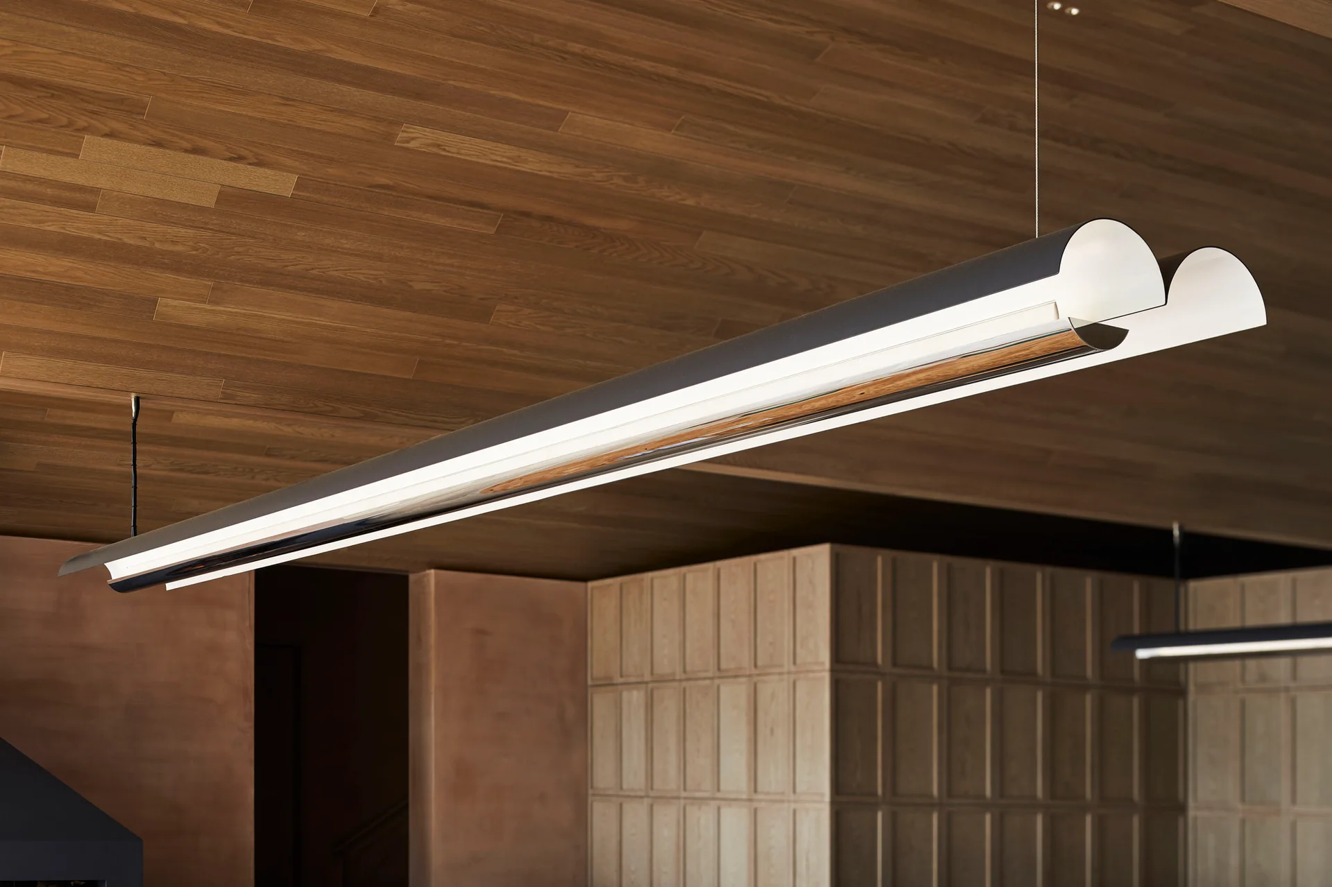 A modern dual-tube linear pendant light hangs from a warm wood plank ceiling above grid-paneled walls.