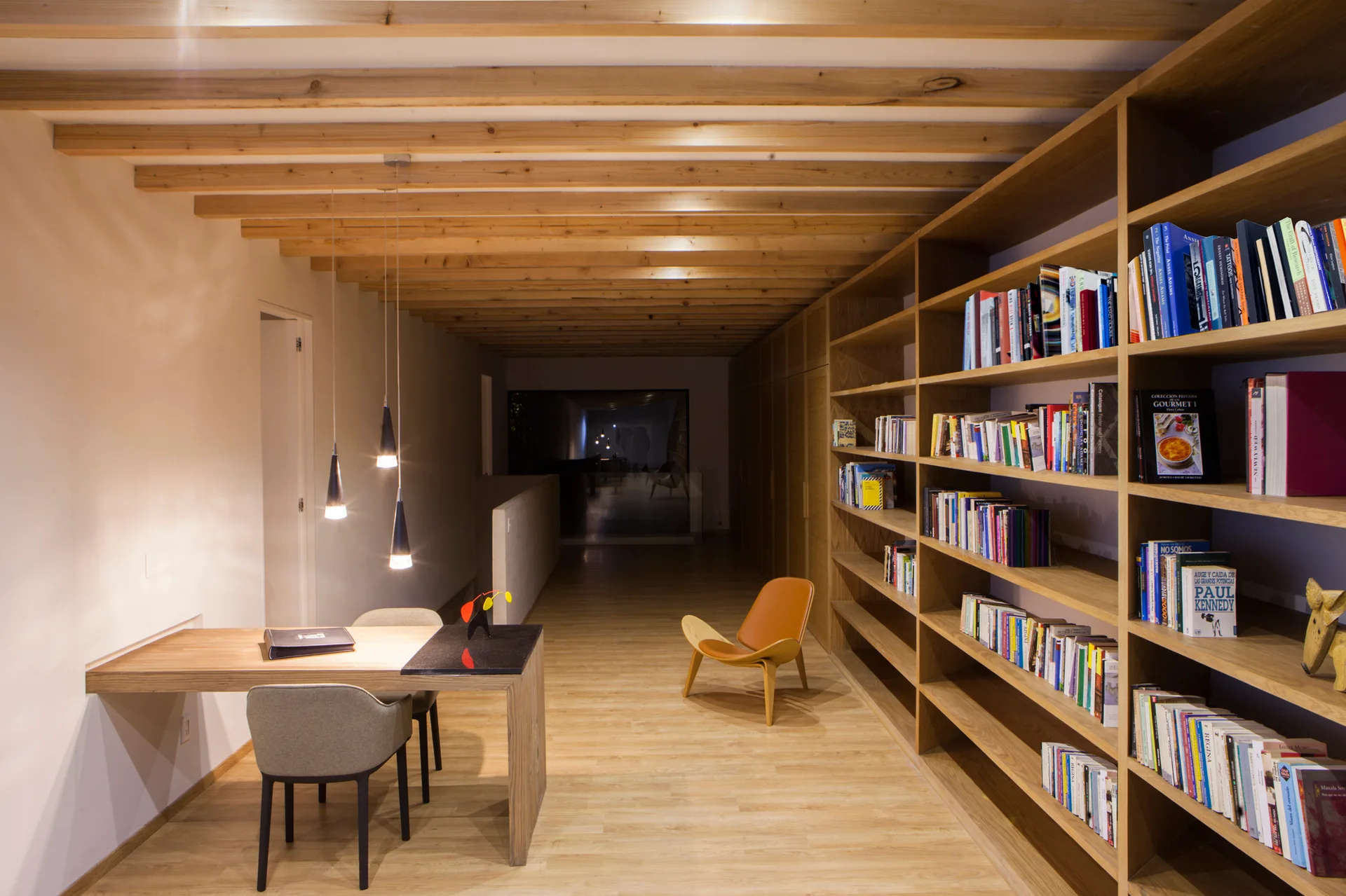 A home library featuring a large wooden bookshelf, a writing desk with pendant lights, and a ceiling with exposed timber beams.