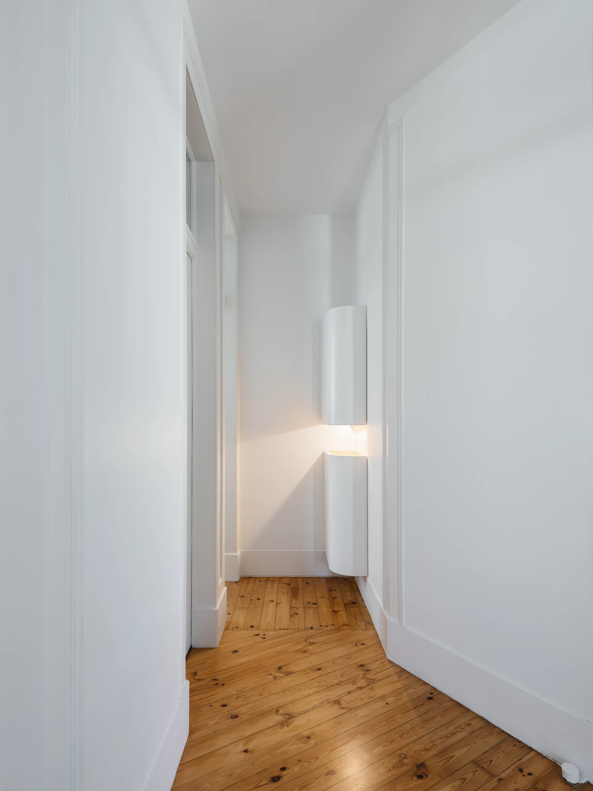 A narrow white hallway featuring diagonal natural wood floorboards and a modern white cylindrical wall-mounted light fixture casting a warm glow.