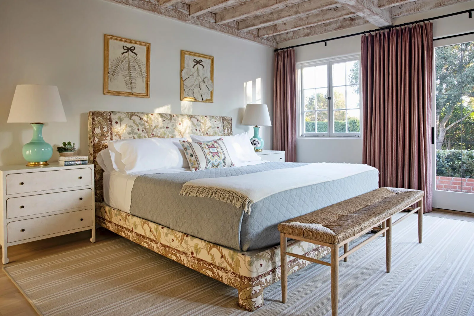 A bedroom featuring an upholstered botanical print bed, matching white nightstands with teal lamps, and whitewashed exposed wood ceiling beams.