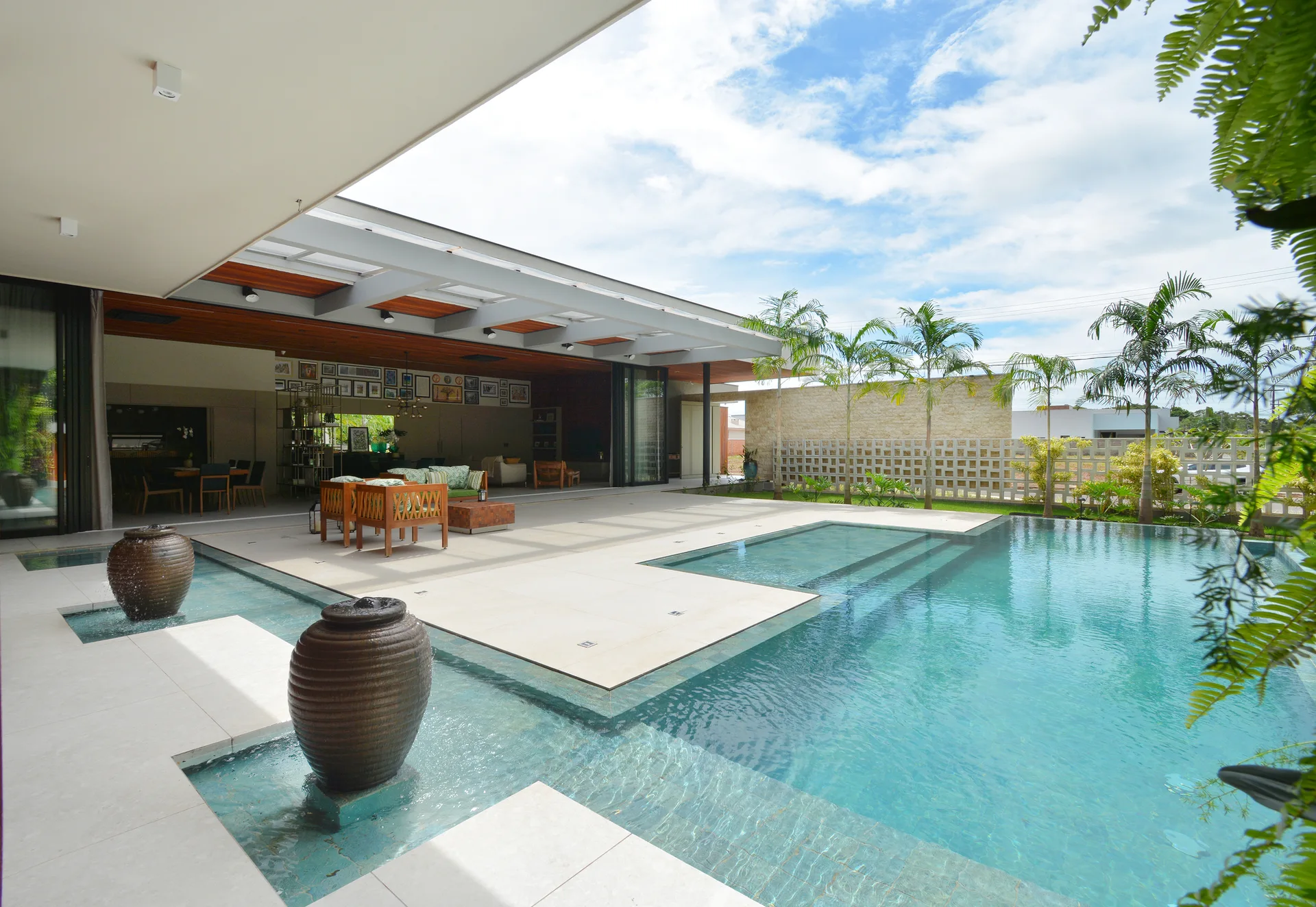 A modern outdoor patio with beige tile flooring sits next to a large blue swimming pool featuring two large ceramic water urn fountains.