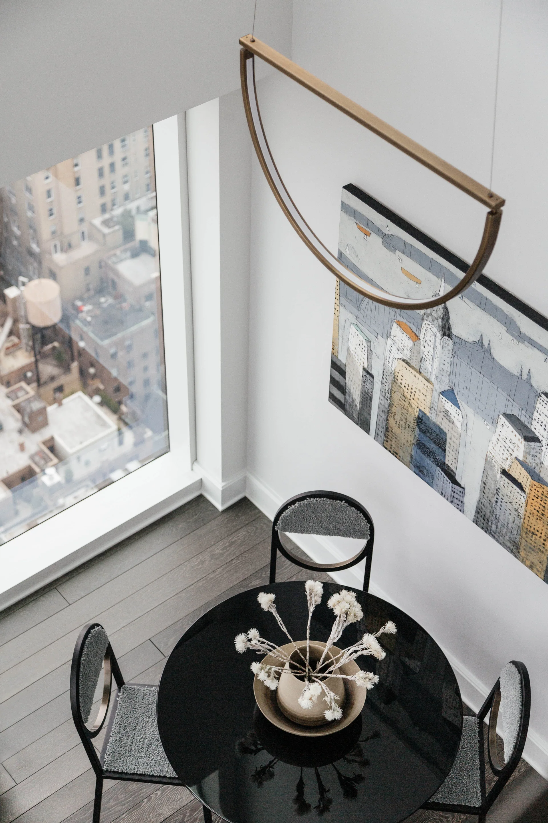 An overhead view of a black round dining table and three grey upholstered chairs beneath a brass semi-circular pendant light next to a window.