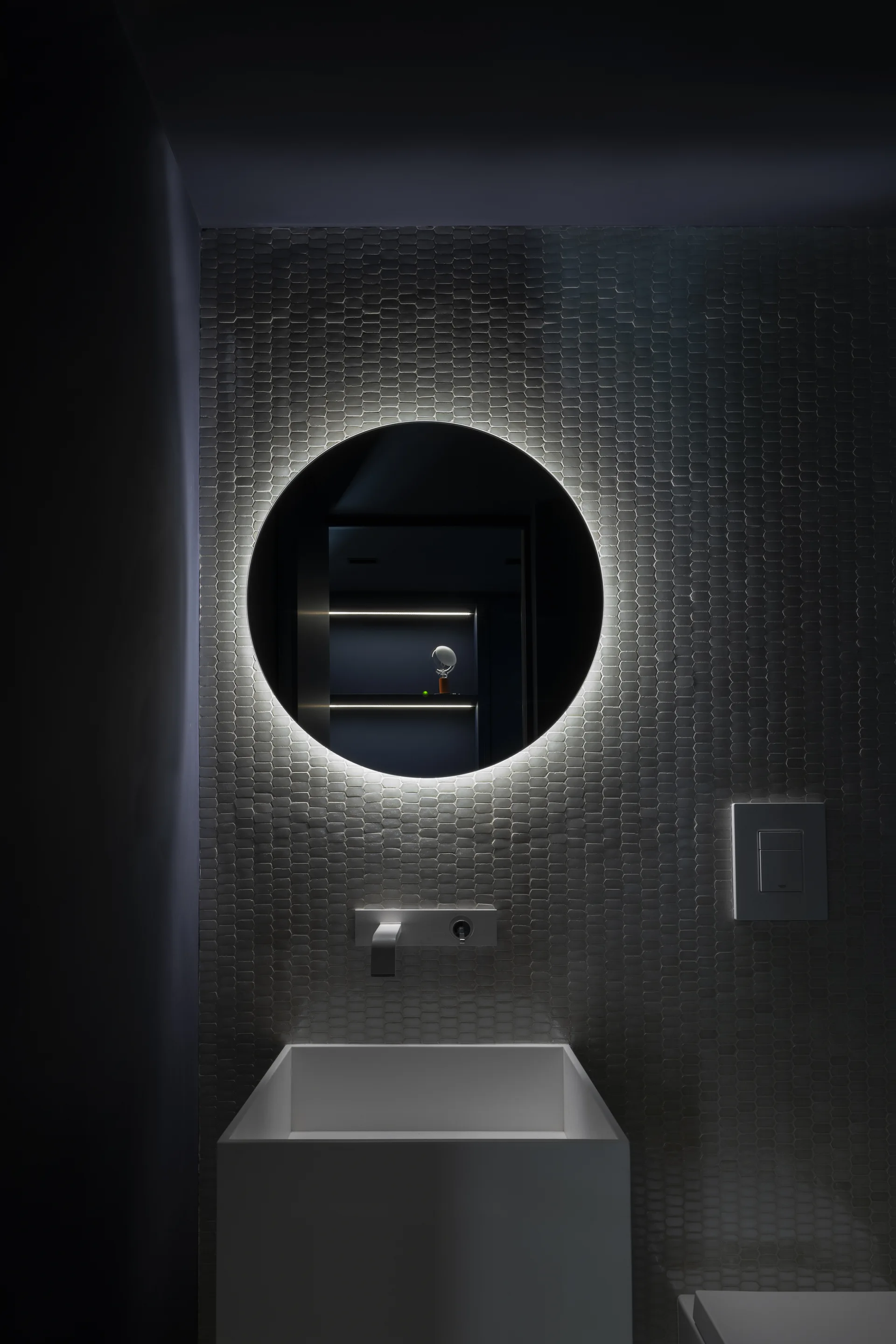 A backlit circular mirror glows on a dark textured mosaic tile wall above a modern white rectangular sink basin with a wall-mounted faucet.