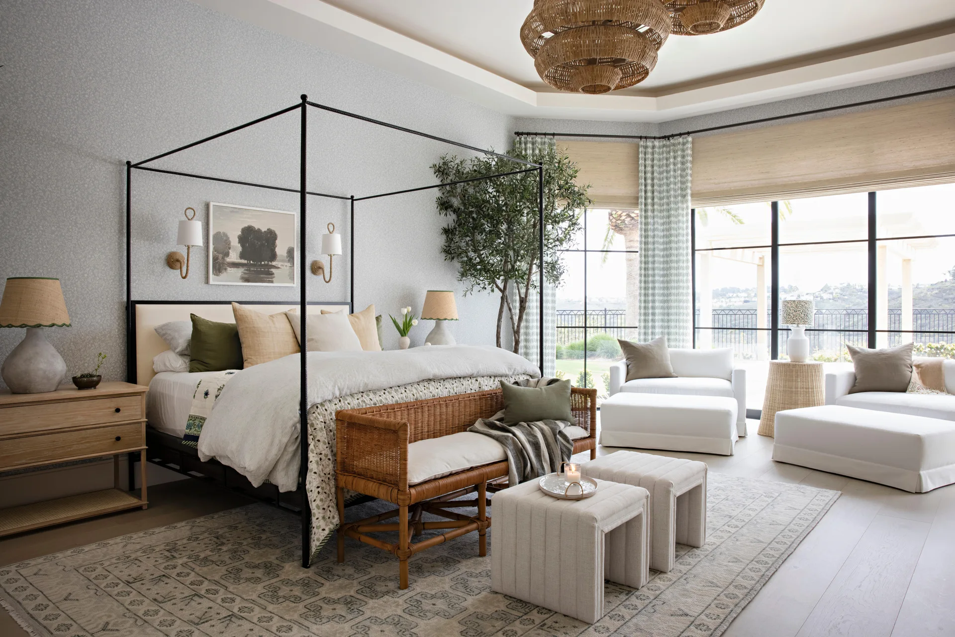 A spacious transitional bedroom featuring a black canopy bed, a woven rattan bench, a patterned rug, and large windows with curtains.