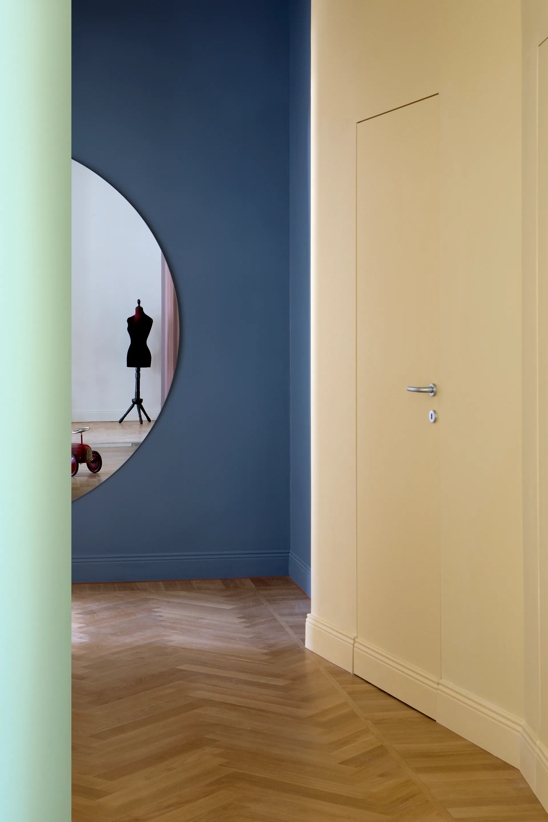 A hallway featuring a flush yellow door, a blue painted wall, a large circular mirror reflecting an adjacent room, and herringbone wood flooring.
