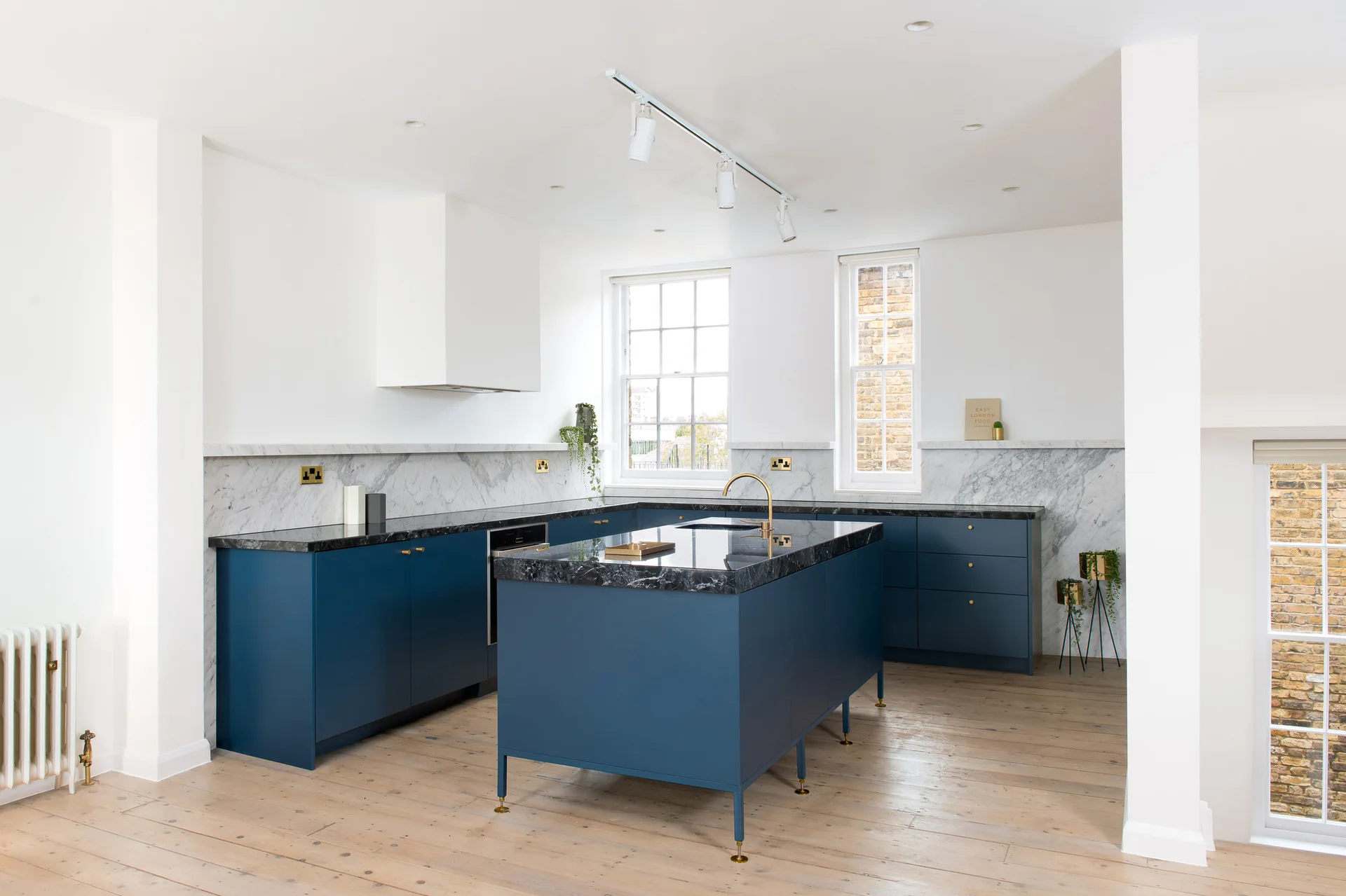 A bright kitchen features matte blue base cabinets, dark marble countertops, a veined marble backsplash, and light wood plank flooring.
