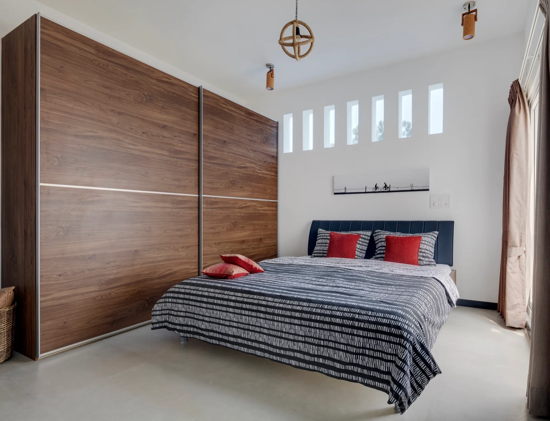 A modern bedroom featuring a large wood-grain sliding door wardrobe, a bed with gray patterned bedding and red pillows, and six high narrow windows.