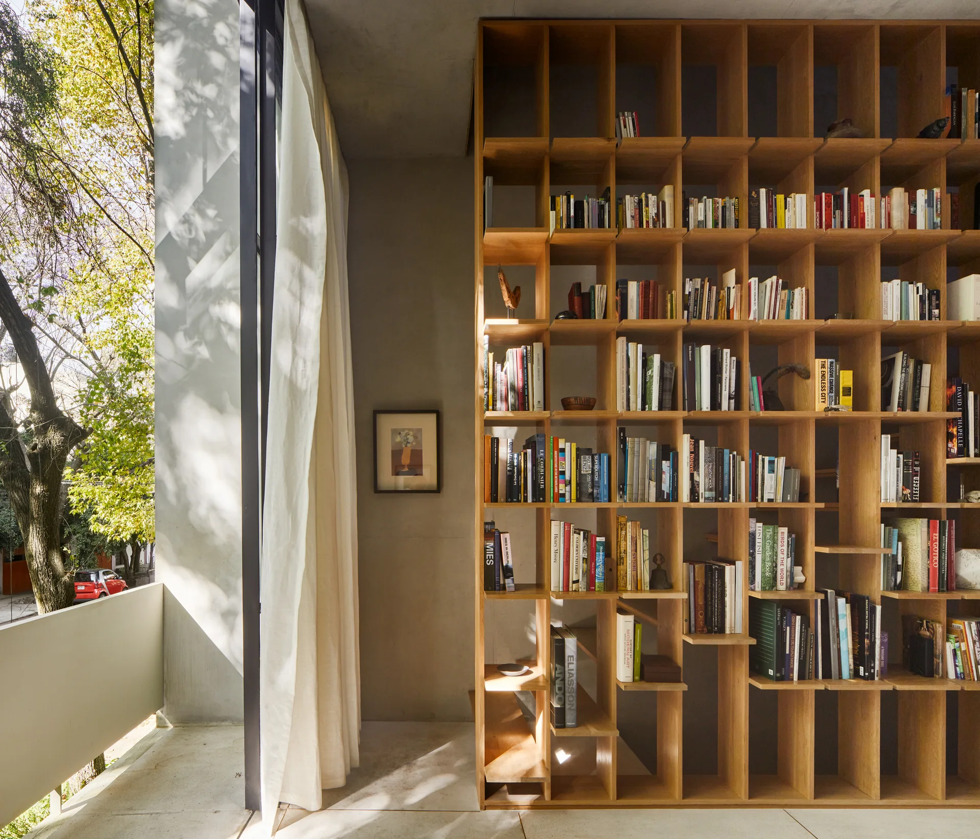 A tall wooden grid bookshelf filled with books stands next to a large window with light curtains overlooking trees.