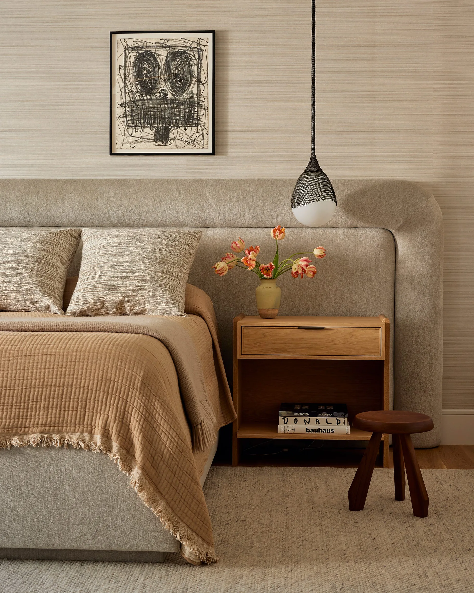 A minimalist bedroom features an upholstered curved headboard, a light wood nightstand with books, a black pendant light, and framed artwork.
