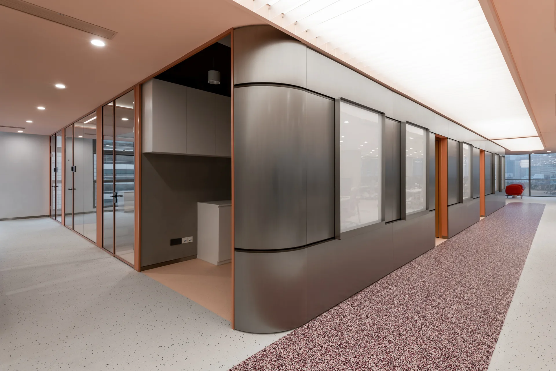 A commercial hallway features a curved brushed metal wall, glass office partitions, and speckled flooring under a translucent ceiling panel.