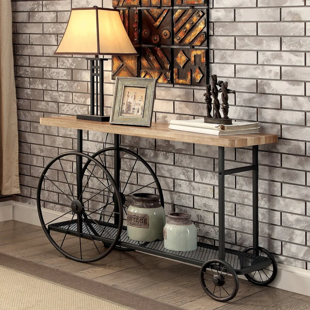A rustic industrial console table with large metal cart wheels features a wood top, a black metal mesh bottom shelf, and sits against a gray brick wall.