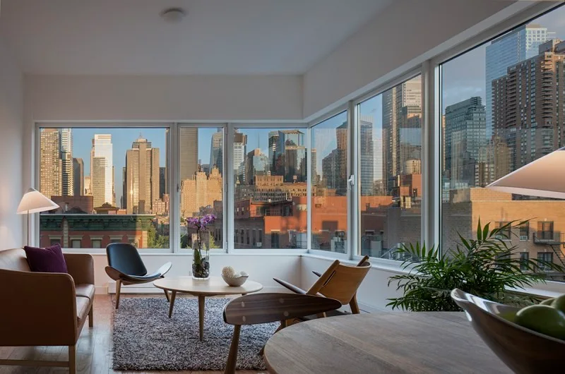 A living room features mid-century modern chairs, a tan leather sofa, a round wooden coffee table, and large corner windows overlooking a city skyline.