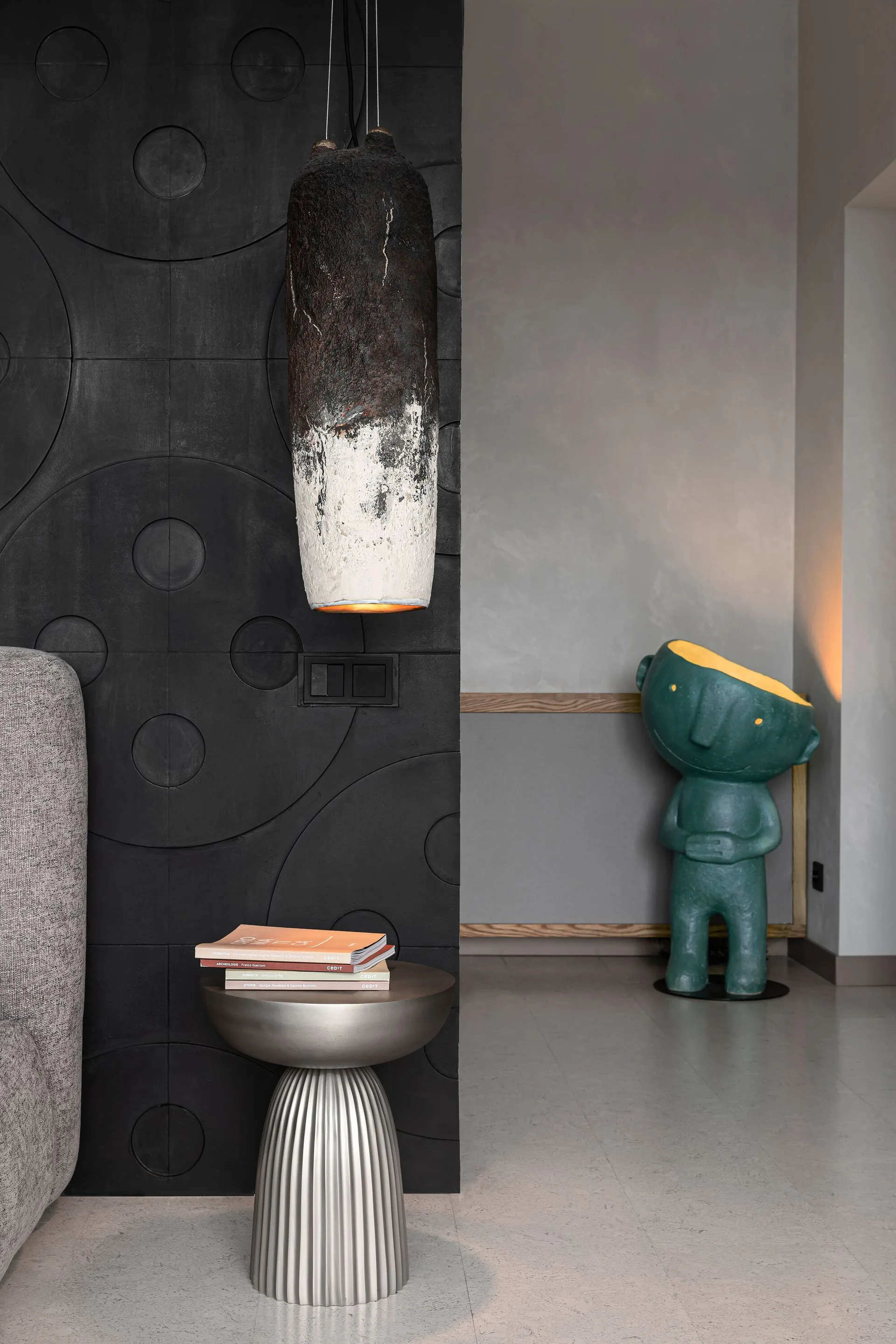 A modern room corner features a black geometric relief accent wall, a fluted metallic side table with books, and a green humanoid floor lamp in the background.