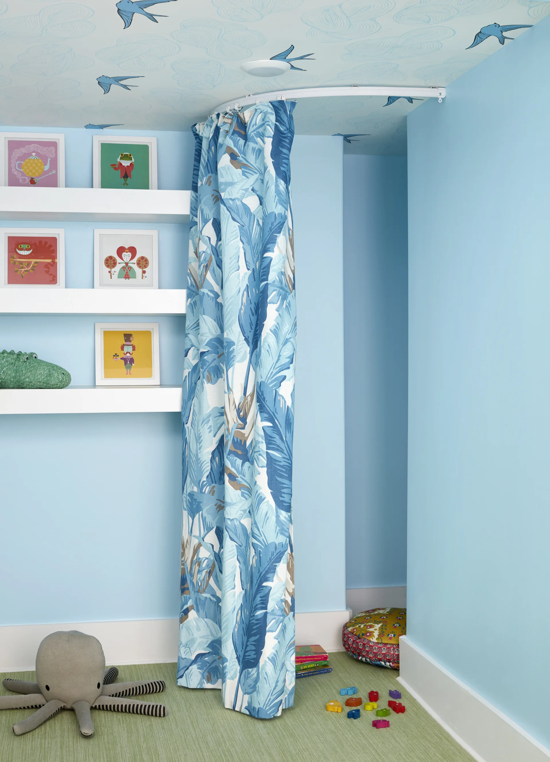 A children's play corner features light blue walls, a curved blue botanical curtain on a ceiling track, white floating shelves, and bird-patterned wallpaper.