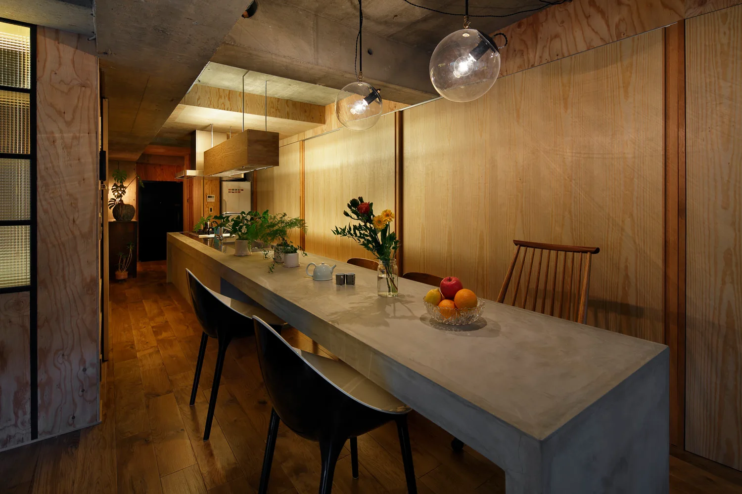 A long concrete kitchen island is surrounded by black chairs and wooden paneling under an exposed concrete ceiling with globe pendant lights.