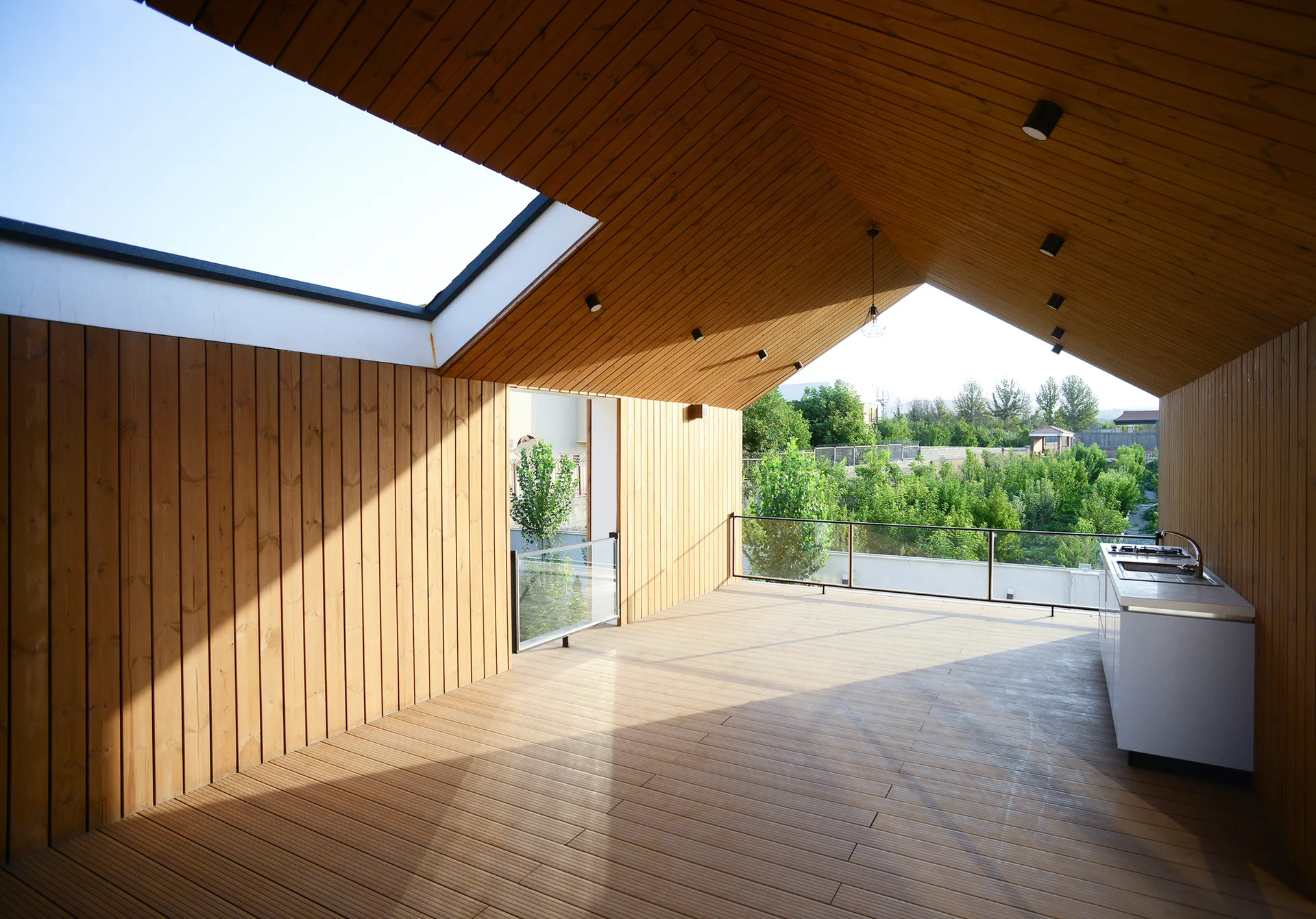 A covered wooden terrace features vertical wood plank walls, an outdoor kitchen cabinet, a glass railing, and a large skylight cut into the vaulted ceiling.