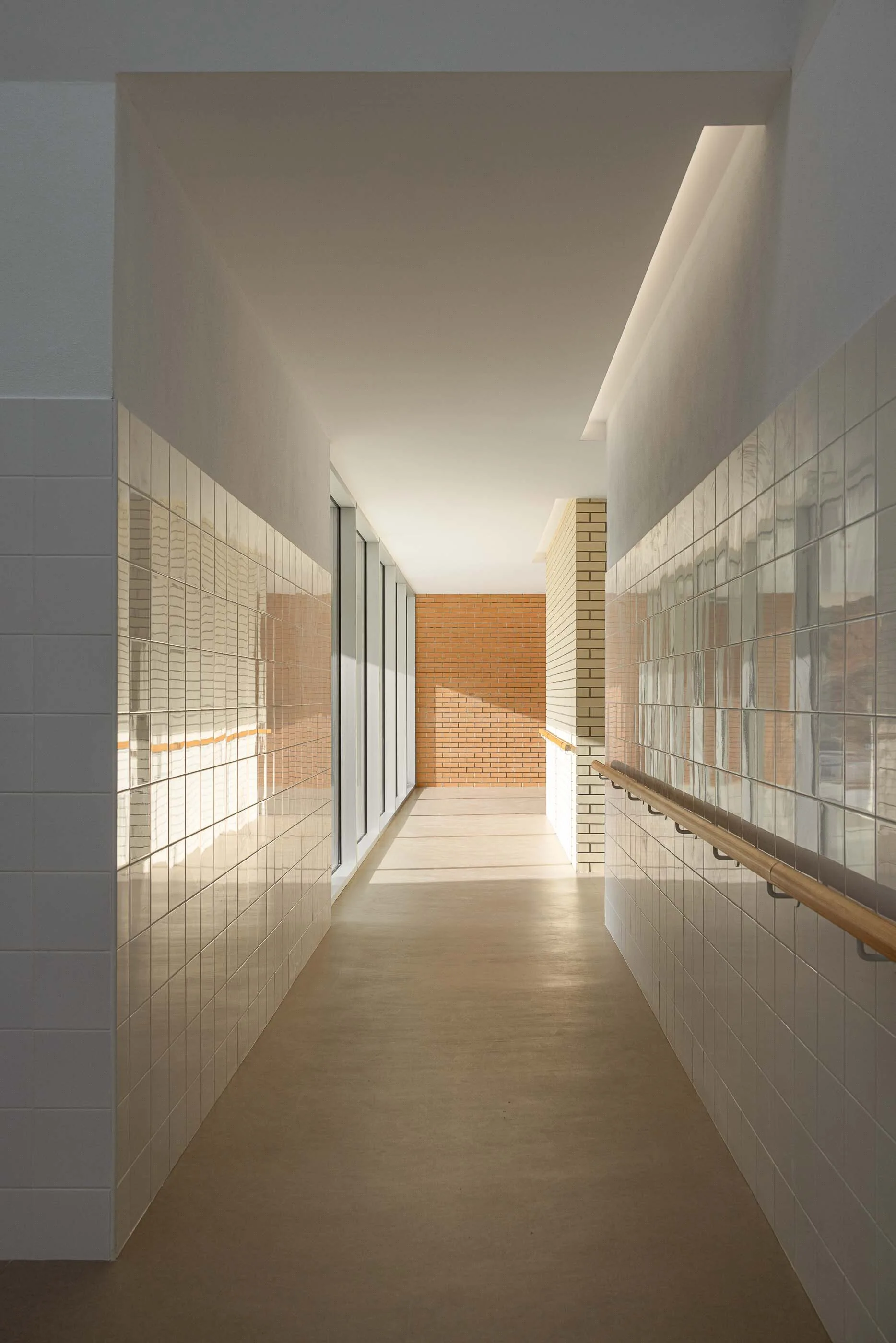 A long interior hallway features glossy white tiled walls with a wooden handrail, leading toward an exposed orange brick wall under bright natural light.