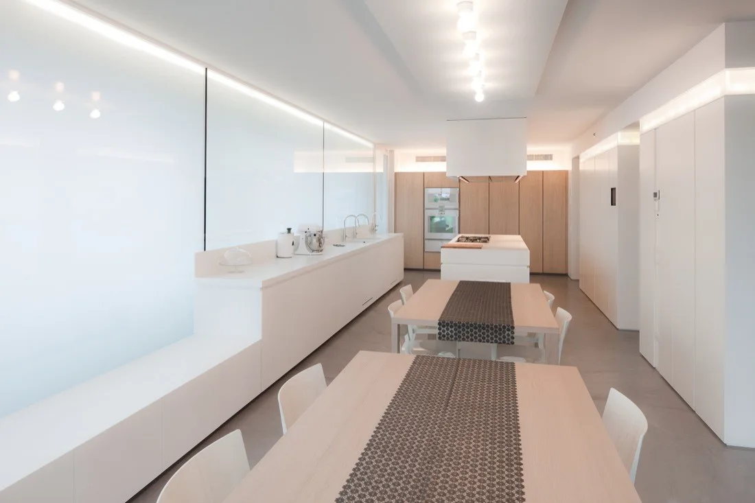 A minimalist white kitchen and dining area features long white handleless cabinetry, linear cove lighting, two wood dining tables, and gray floors.
